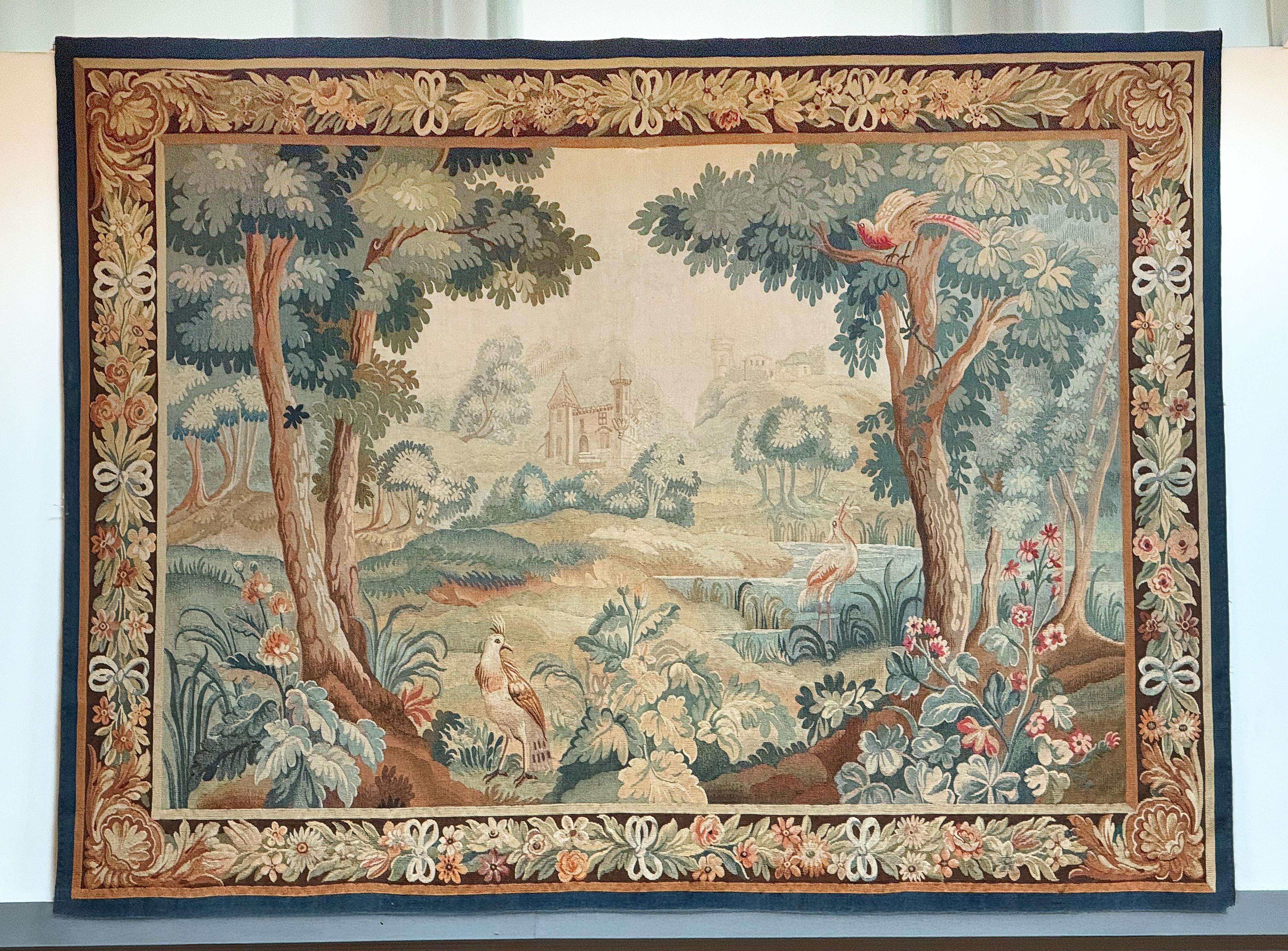 A fine large French Aubusson style tapestry from the 19th century, featuring a verdure landscape scene with colorful birds in the foreground, and a castle in the background surrounded by a lake and woodland. 

The tapestry is framed by an ornate