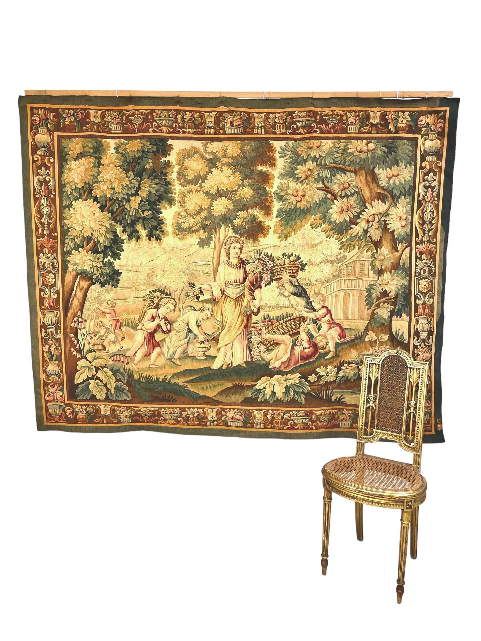 A fine Aubusson tapestry depicting a robed lady and several mischievous children bearing baskets of fruit, in a typically rural and architectural-themed landscape. A wide border on all four sides is densely decorated with flower-filled urns and