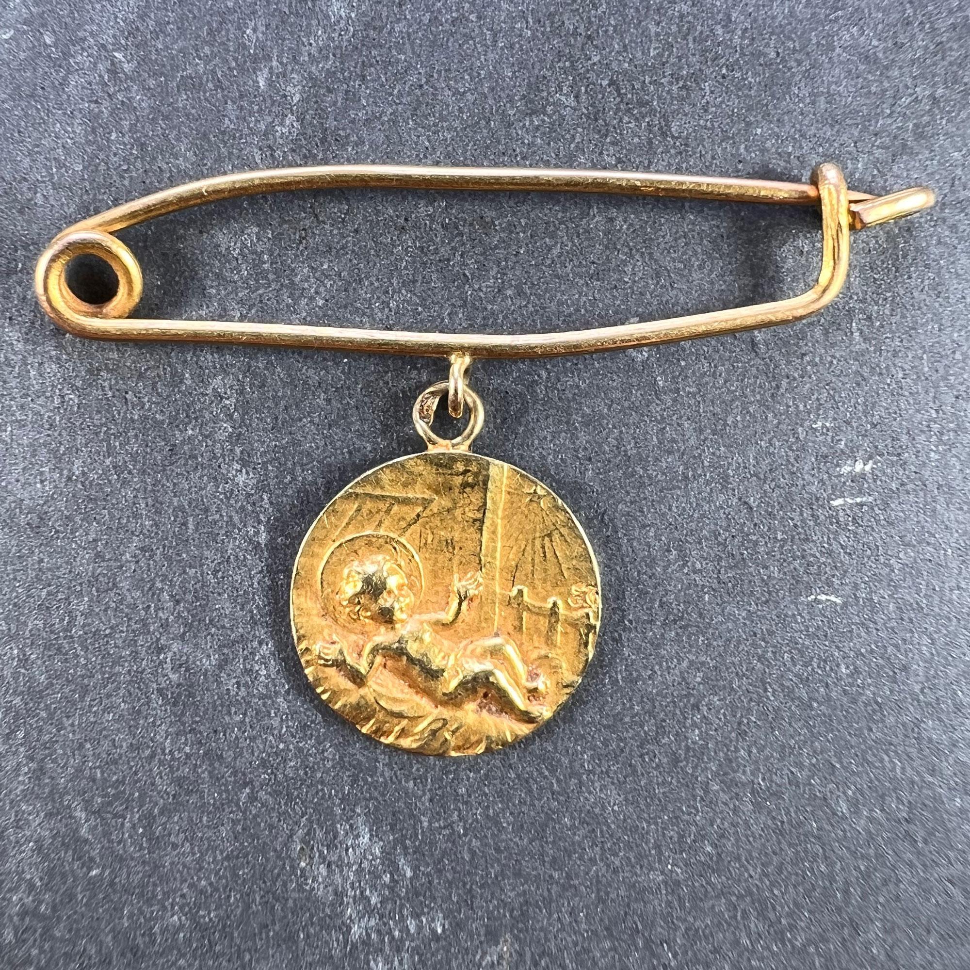 An 18 karat (18K) yellow gold charm pendant brooch of a medal depicting a baby in an infant’s crib with rays of light streaming down onto it from a star, suspended from a yellow gold nappy pin or safety pin. Stamped with the eagle’s head for French