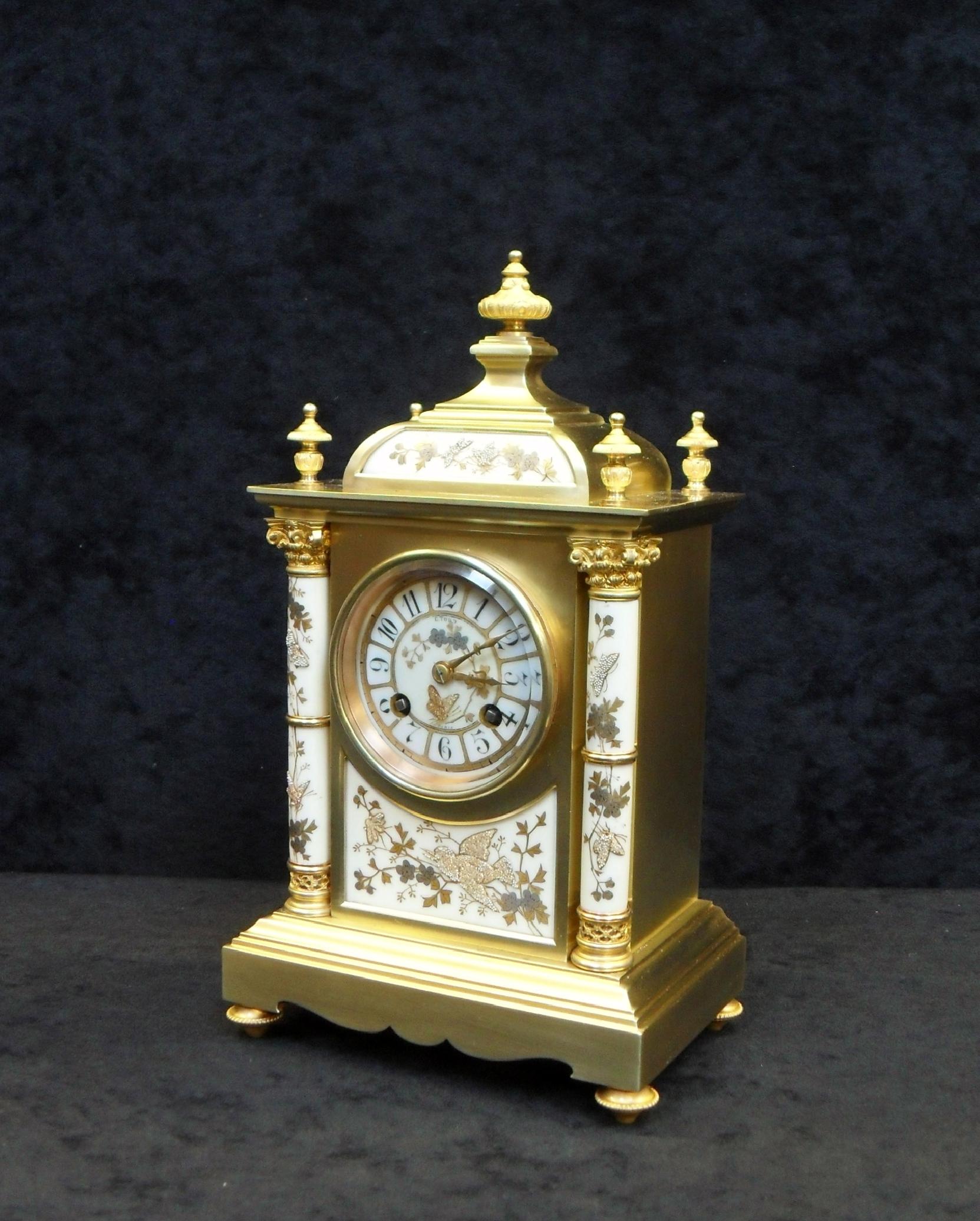 A decorative French Belle Époque brass mantel clock with inset porcelain panels, dial and columns finished with brass capitals and finials to the top. The porcelain has a hand painted silver and gilt floral leaf design with bird and butterfly glass
