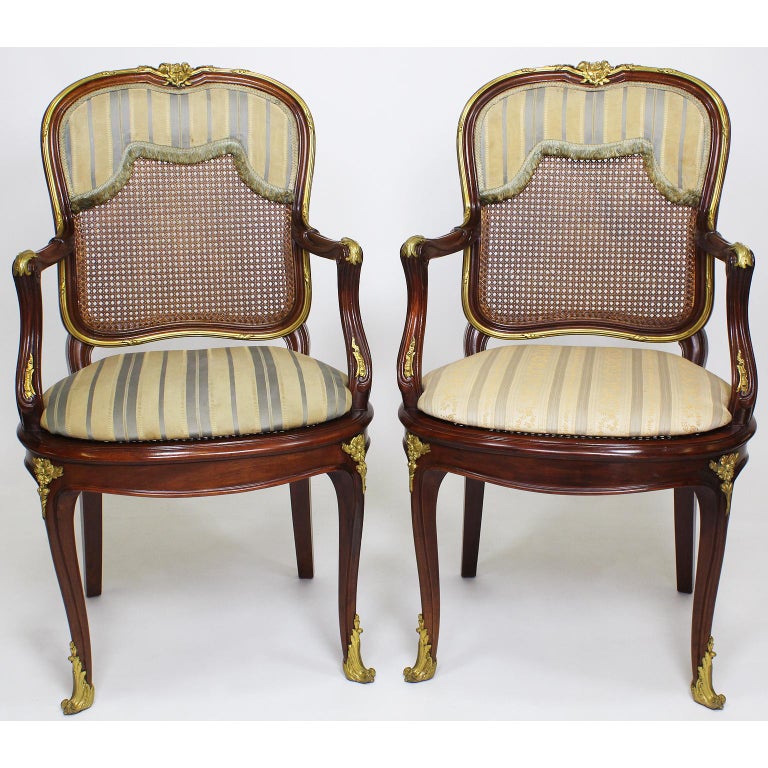 A fine set of fourteen, French, 19th-20th century Belle Époque Louis XV style mahogany and ormolu-mounted dining chairs, comprising of twelve chairs and two armchairs, all with cane backs and seats and padded flat pillows, with floral and banded