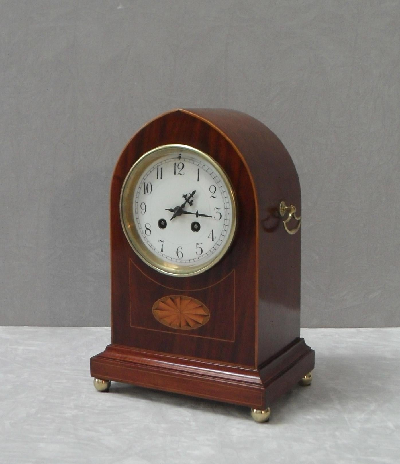 A good quality Belle Époque figured mahogany lancet top mantel clock with fan inlay and boxwood stringing to the front of the case with brass carrying handles to the side stood on brass ball feet. The clock has a white enamel dial with a French