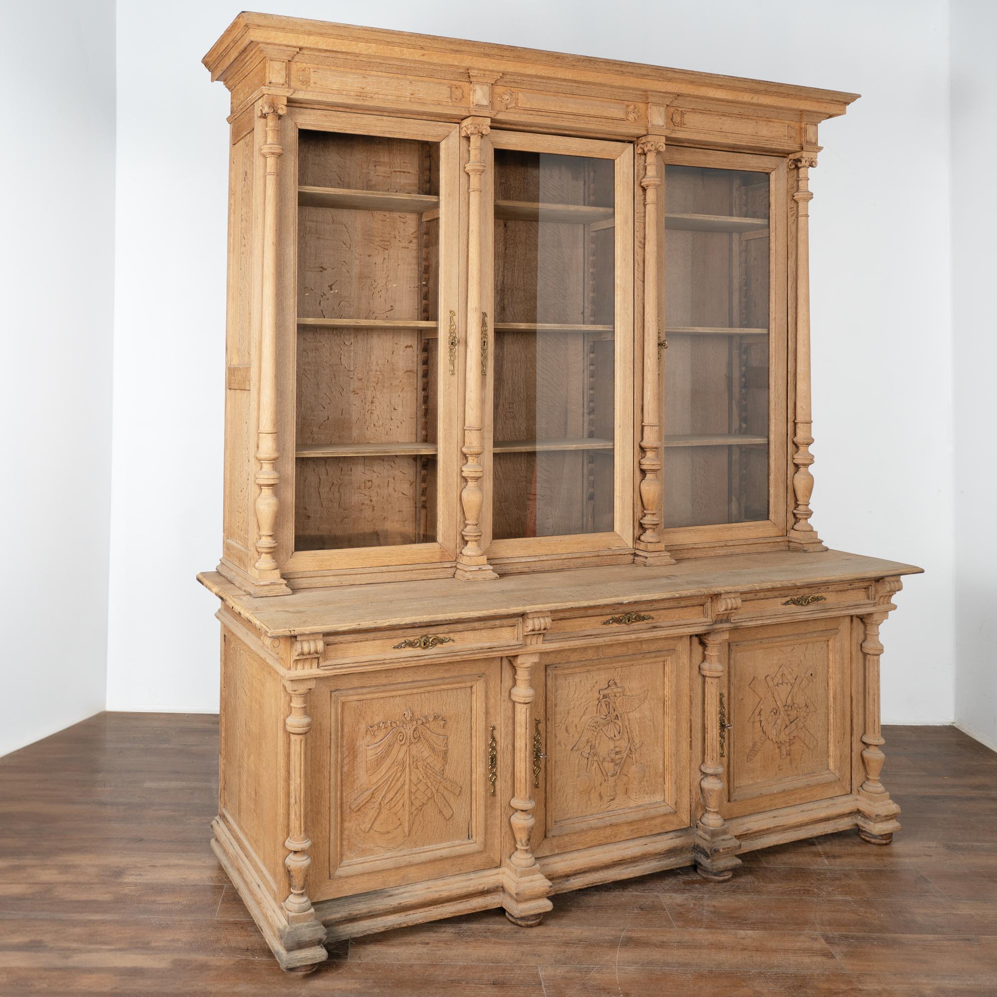 This large bleached oak bookcase is statuesque in size and is impressive with turned columns and 3 carved panel doors.
At 8' tall, the glass door panes add to its visual impact displaying any collection held within; adjustable shelving adds to the