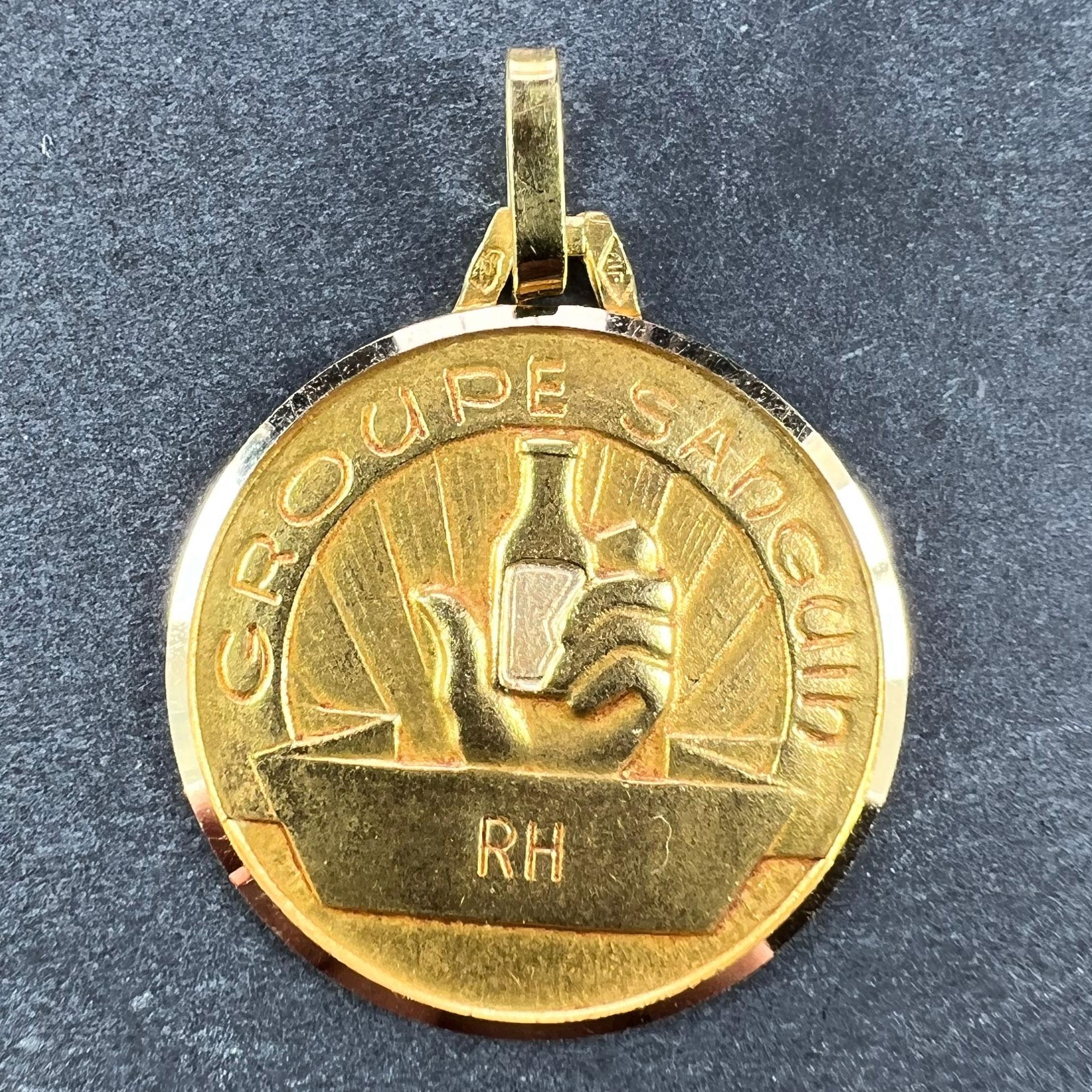 A French 18 karat (18K) yellow gold charm pendant designed as a medal depicting a hand holding a bottle aloft in the rays of a rising or setting sun, with the words 'Groupe Sanguin' above and around it, with RH on a banner below with space for