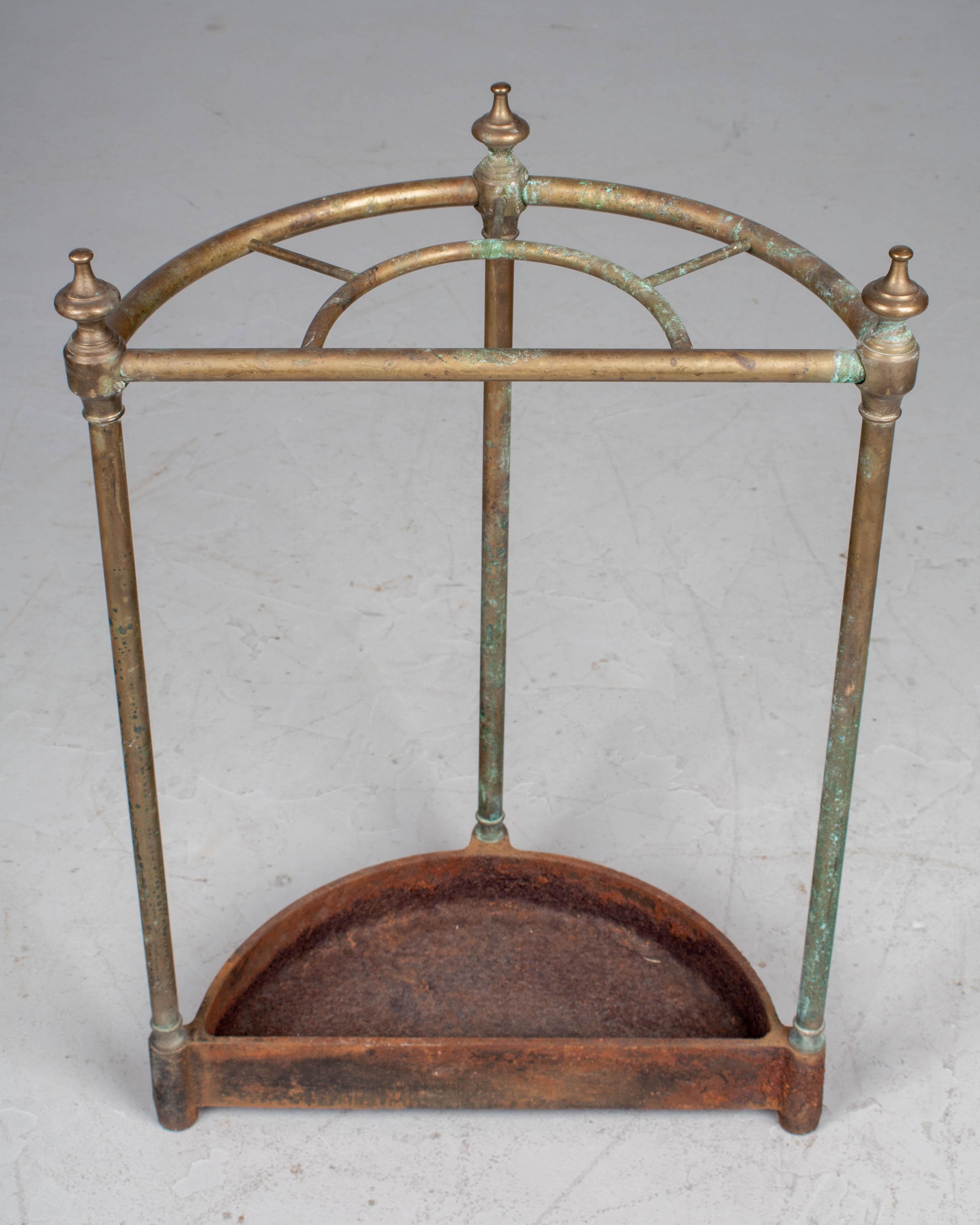 A French brass and cast iron umbrella stand. Demilune form with brass tubing and decorative finials. Cast iron drip tray base is heavily rusted with remnants of black showing through. Beautiful old patina. 
More photos available upon request. We