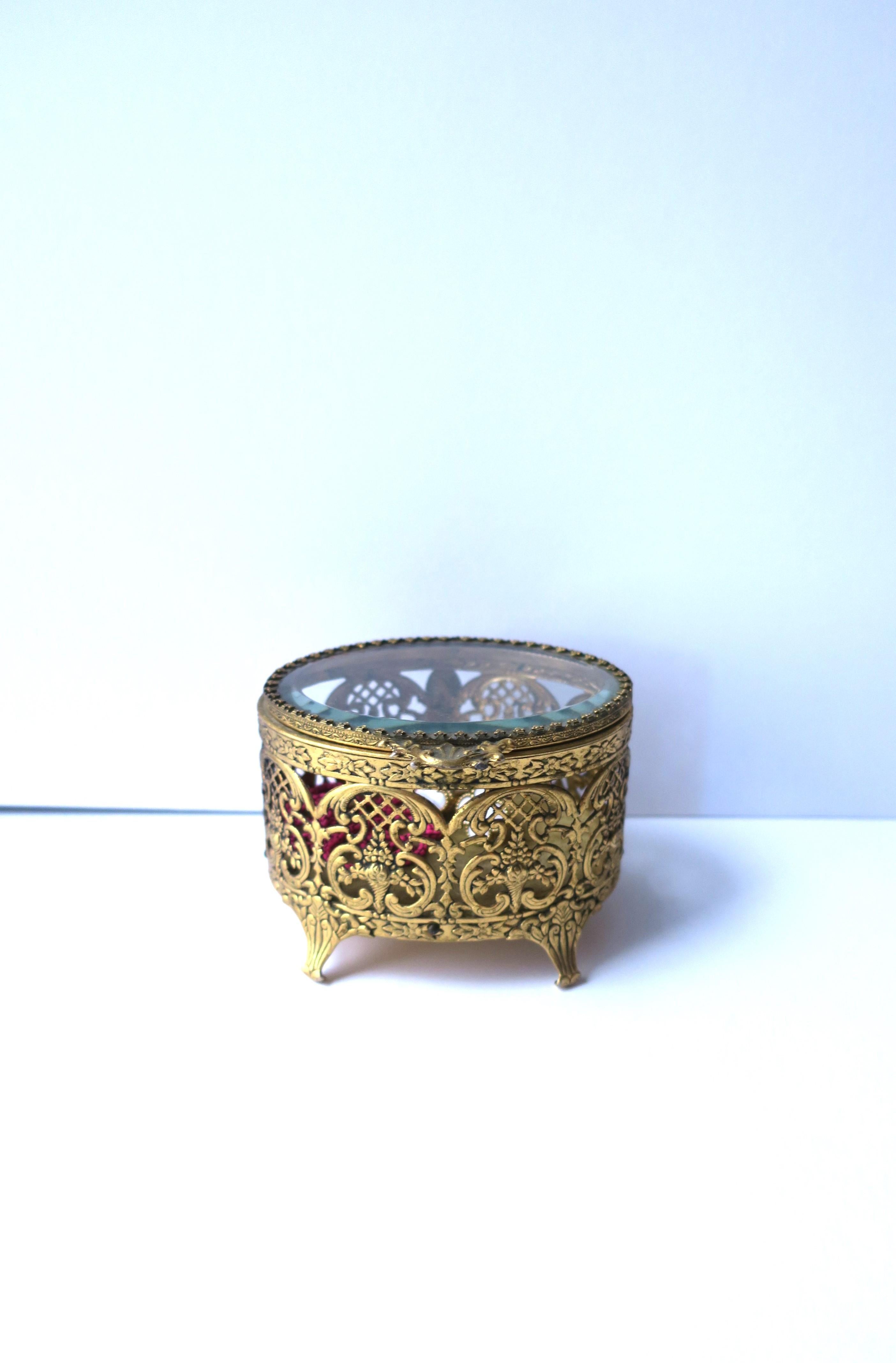 A French brass and glass jewelry box, in the Rococo style, circa early-20th century, France. This oval jewelry box is brass with a filigree design, a beveled glass hinged top, cotton velvet interior, and small curved feet. Piece may work well for a