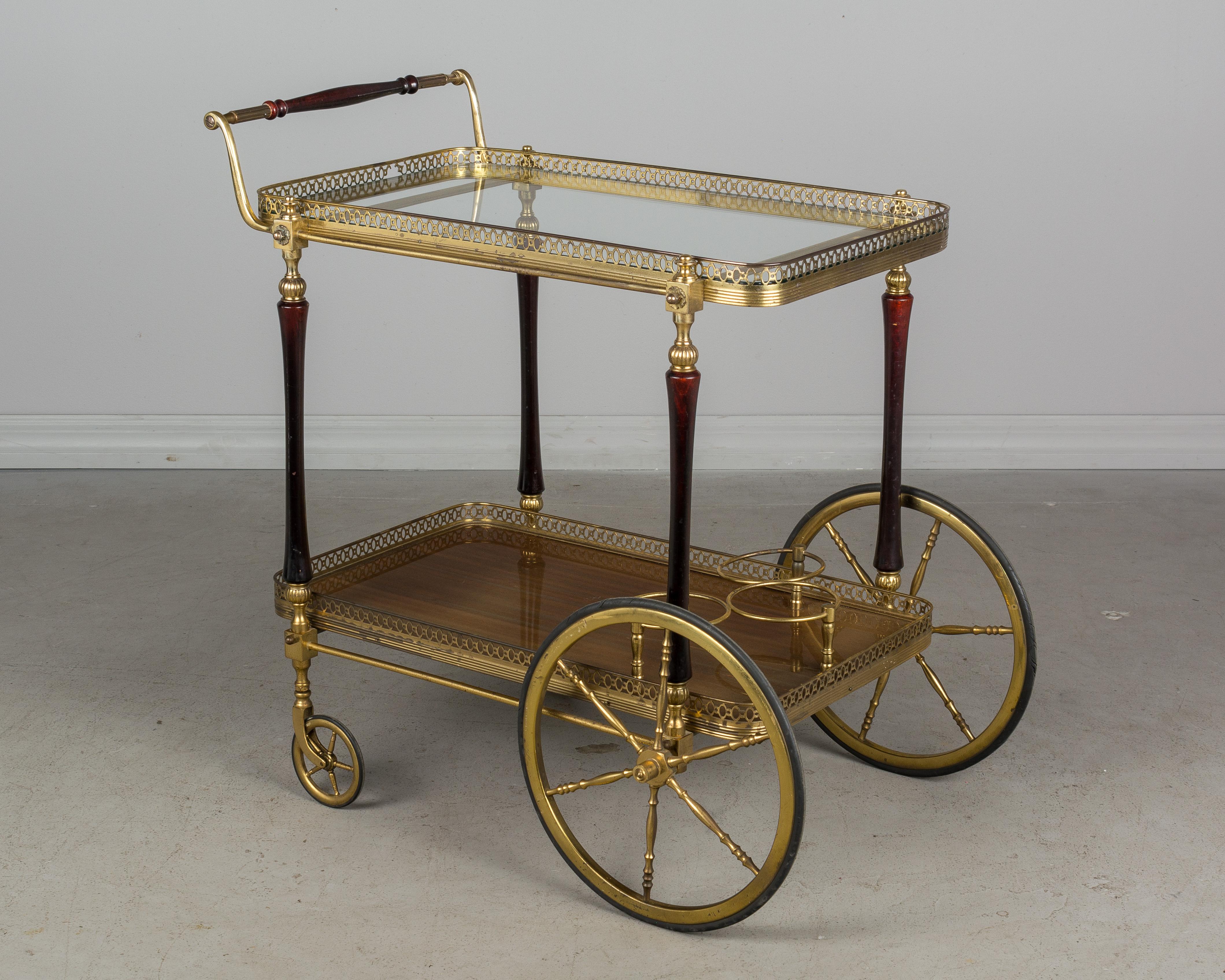 A 1940s French solid brass and mahogany bar cart with glass shelves surrounded by a decorative brass gallery. The brass has not been polished and retains and old warm patina with pitting in places. The glass of the upper shelf is removable and has a