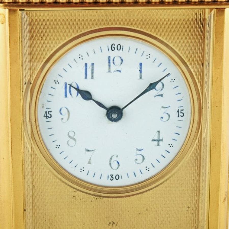 A late 19th to early 20th century brass carriage clock timepiece.

The clock has a white dial with blue and black numerals surrounded by an engine turned brass frame.

The clock has short bracket feet and a hinged carrying handle.

The clock