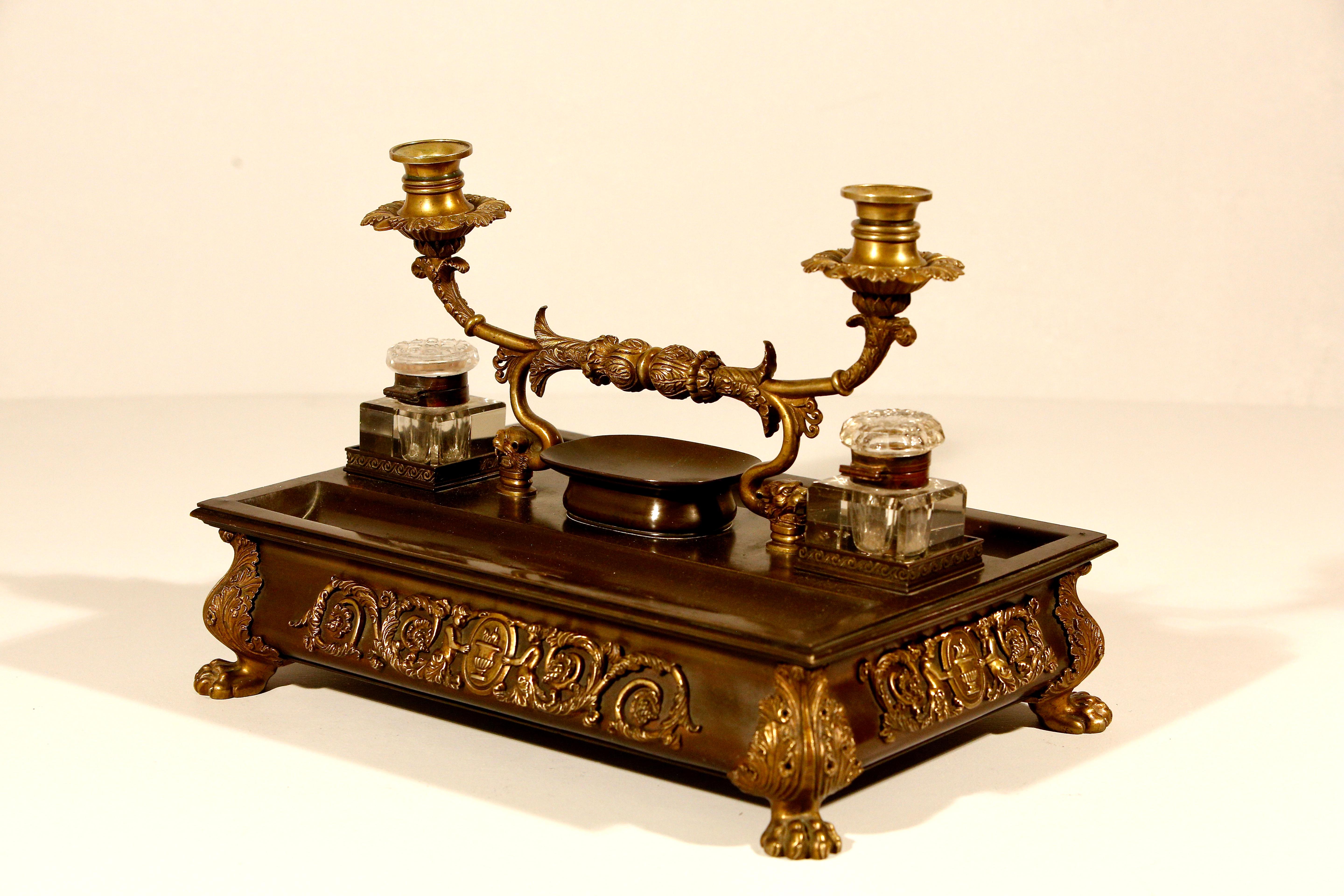 A large and impressive early 19c French bronze and gilt bronze inkwell, the central chased handle with a pair of candle arms terminating in lion masks, 2 tray wells either side of a raised dish with 2 cut glass inkwells supported on a shaped bronze
