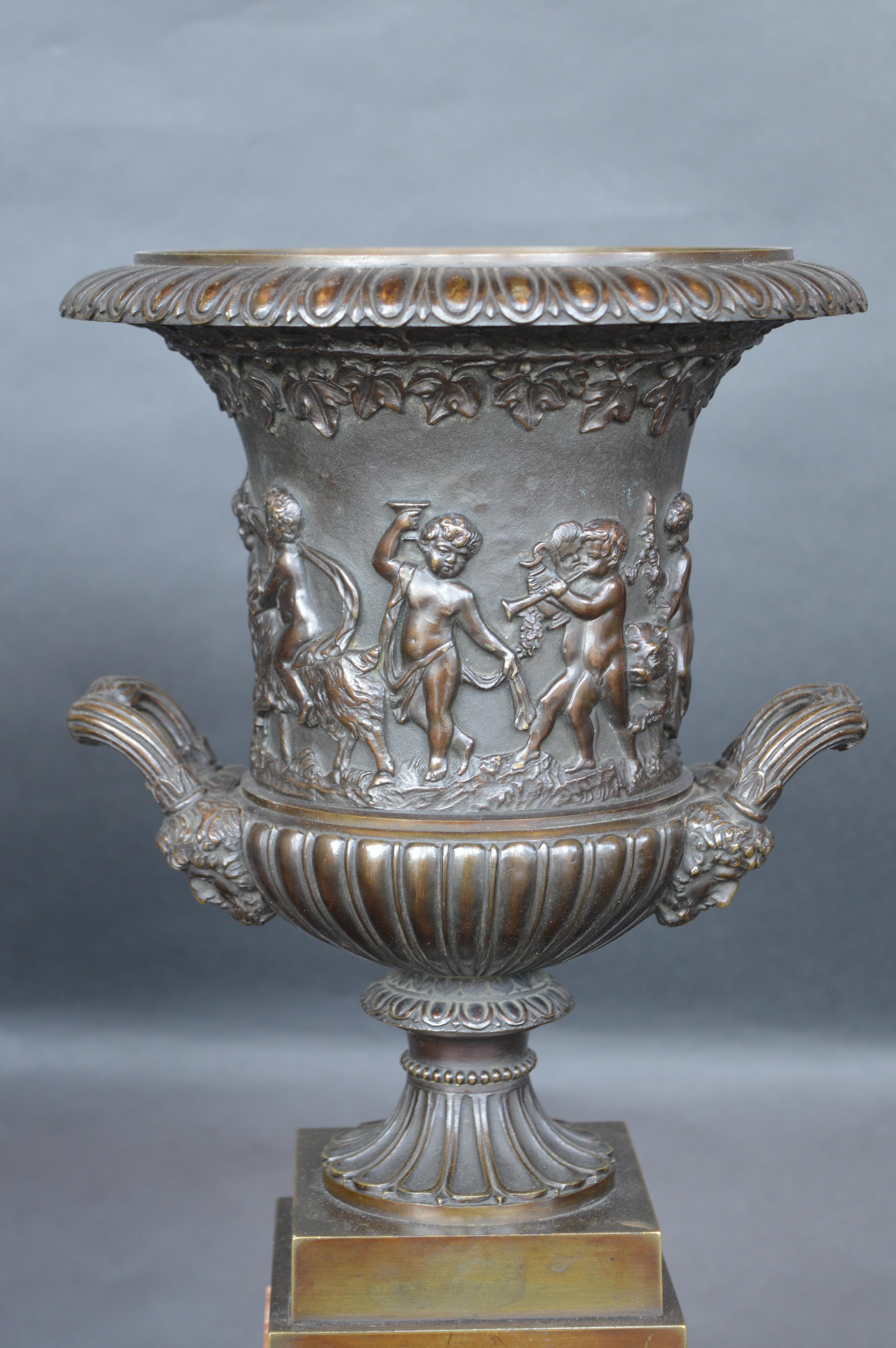 Bronze patinated vases sit on top of a sienna marble base. Cherubs surround the vase, feeding grapes to a woman in a chariot.