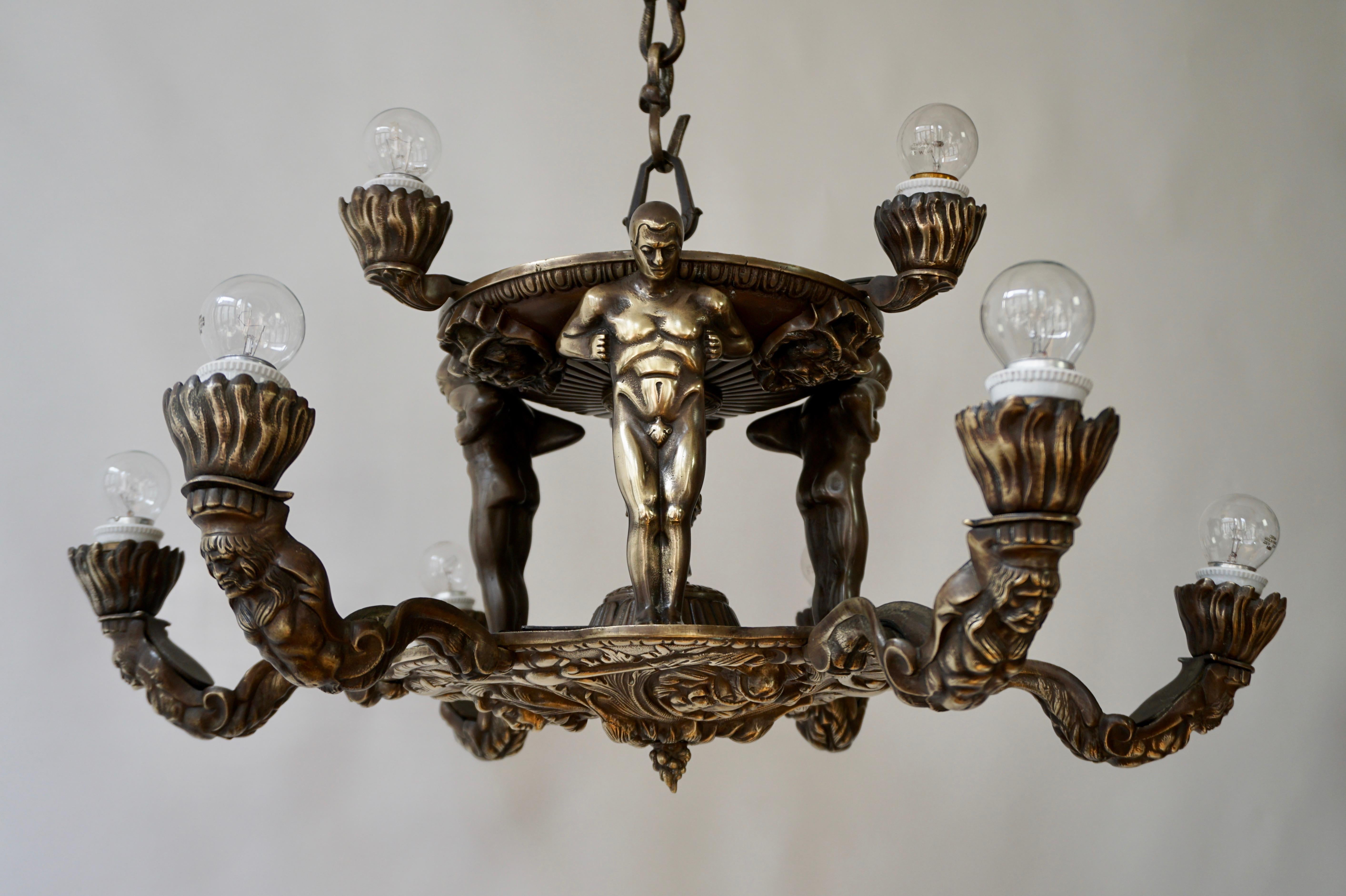 A remarkable two tier bronze six light French Hollywood Regency chandelier, the upper part inspired by Wiener Werkstätte Art Deco designs showing three male nude figures supporting a fountainlike structure, the lower part decorated with medallions