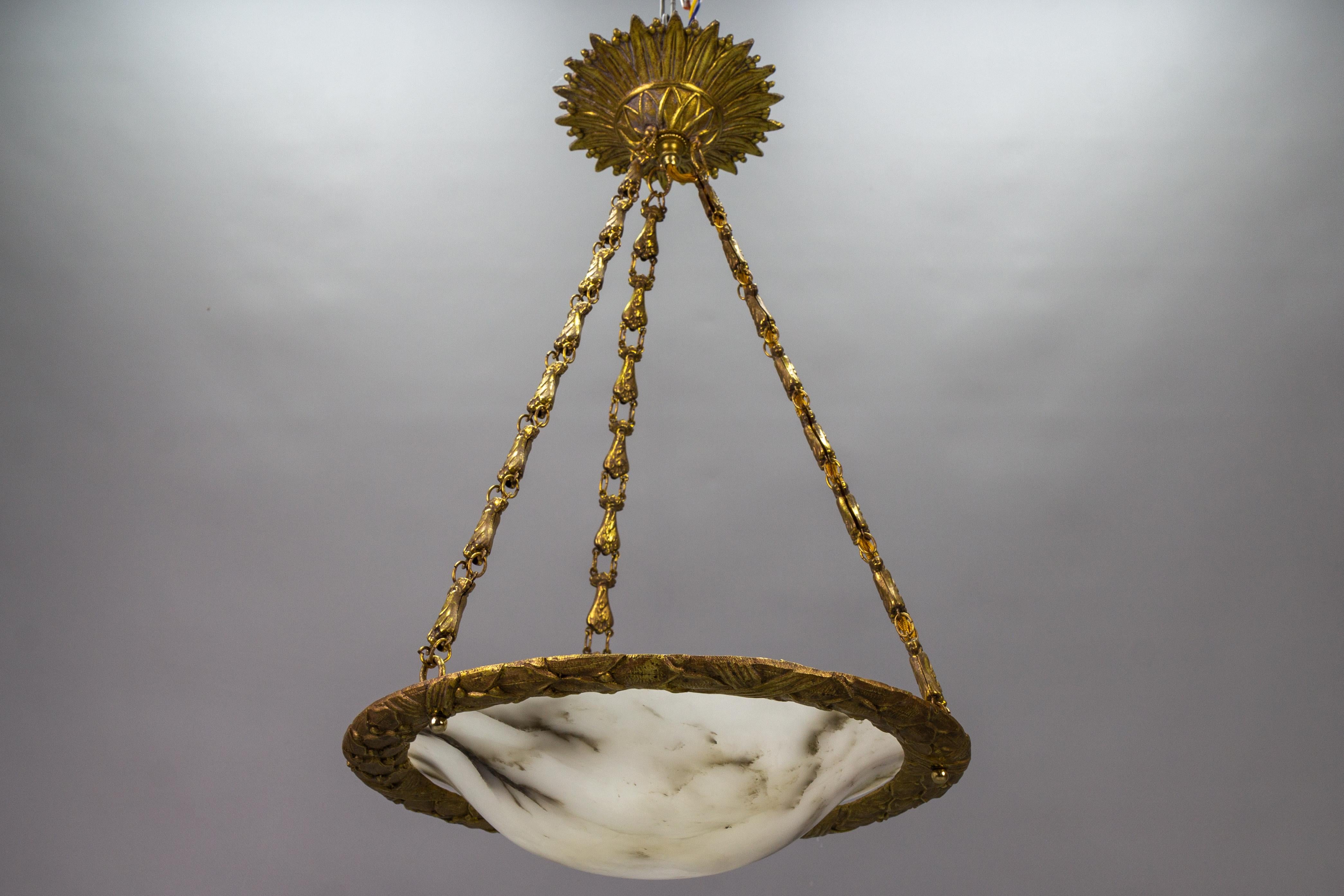 Antique French bronze, brass, and alabaster pendant light from ca. 1920.
This adorable Art Deco French pendant light features a soft white alabaster lampshade - bowl with dark veins in an ornate bronze rim, hung on three decorative brass chains.