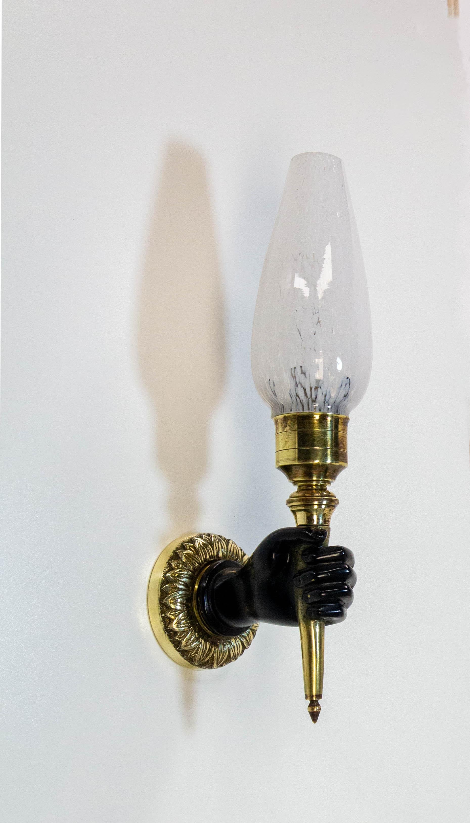 1950s wall lamp consisting of a frieze of flowers (Raie-de-coeur) supporting a Nubian hand, which holds a torch.
The white speckled glass, also known as Clichy glass, is not the original, but its elegance perfectly suits the style of this