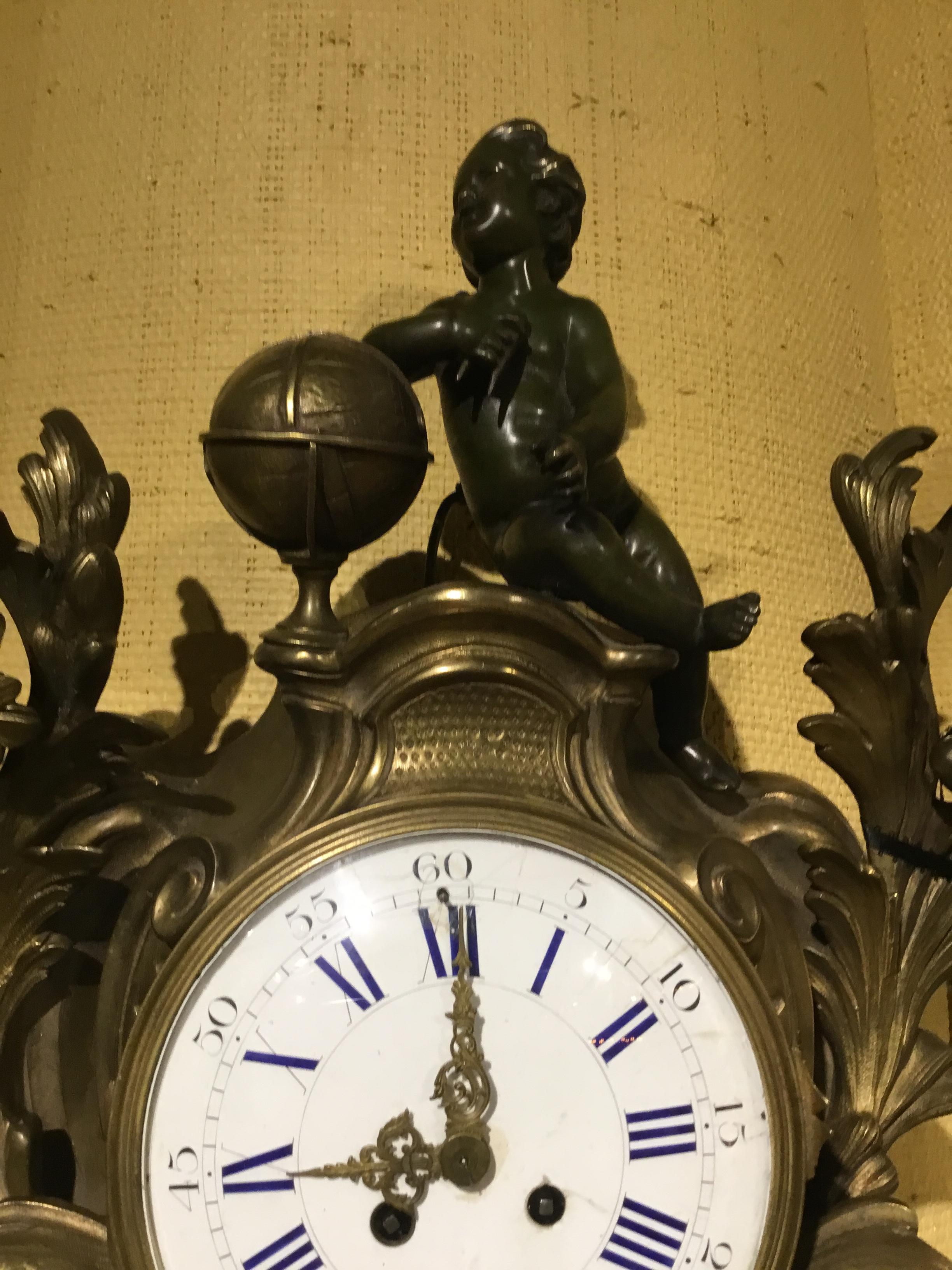 Large cartel clock in bronze with a patinated putto that sits at the crest of this exceptional clock.
Flourishes and foliate designs grace the sides of the circular clock face that is made of white
Enamel with blue Roman numerals. The pendulum and