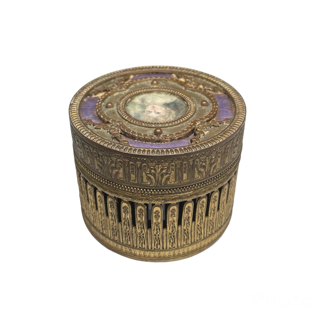 This is a French patinated bronze metal round trinket lidded box. It depicts a pierced round metal box decorated with a relief of foliages. The lid is adorned in the center with a hand painted portrait of a young girl enframed with a quatrefoil.