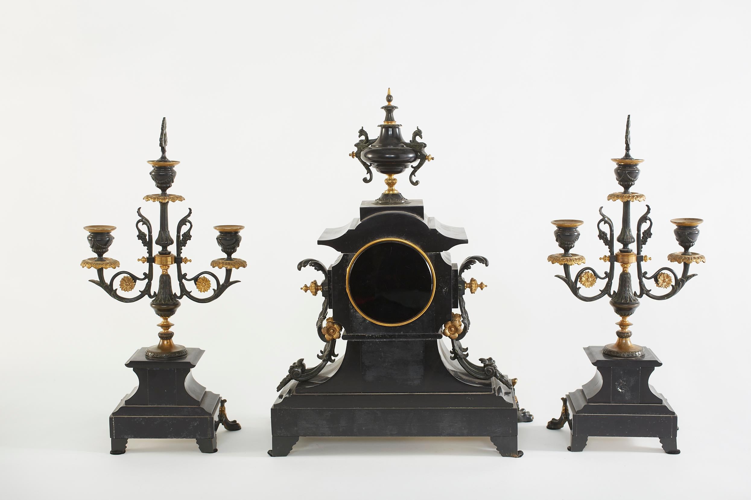 French bronze & polish black marble with applied bronze trim three piece clock garniture set. The clock surmounted by an urn flanked & griffins. Clock face with gold Roman numerals and spade hands. Illegible signature, brass 8 day movement with rack