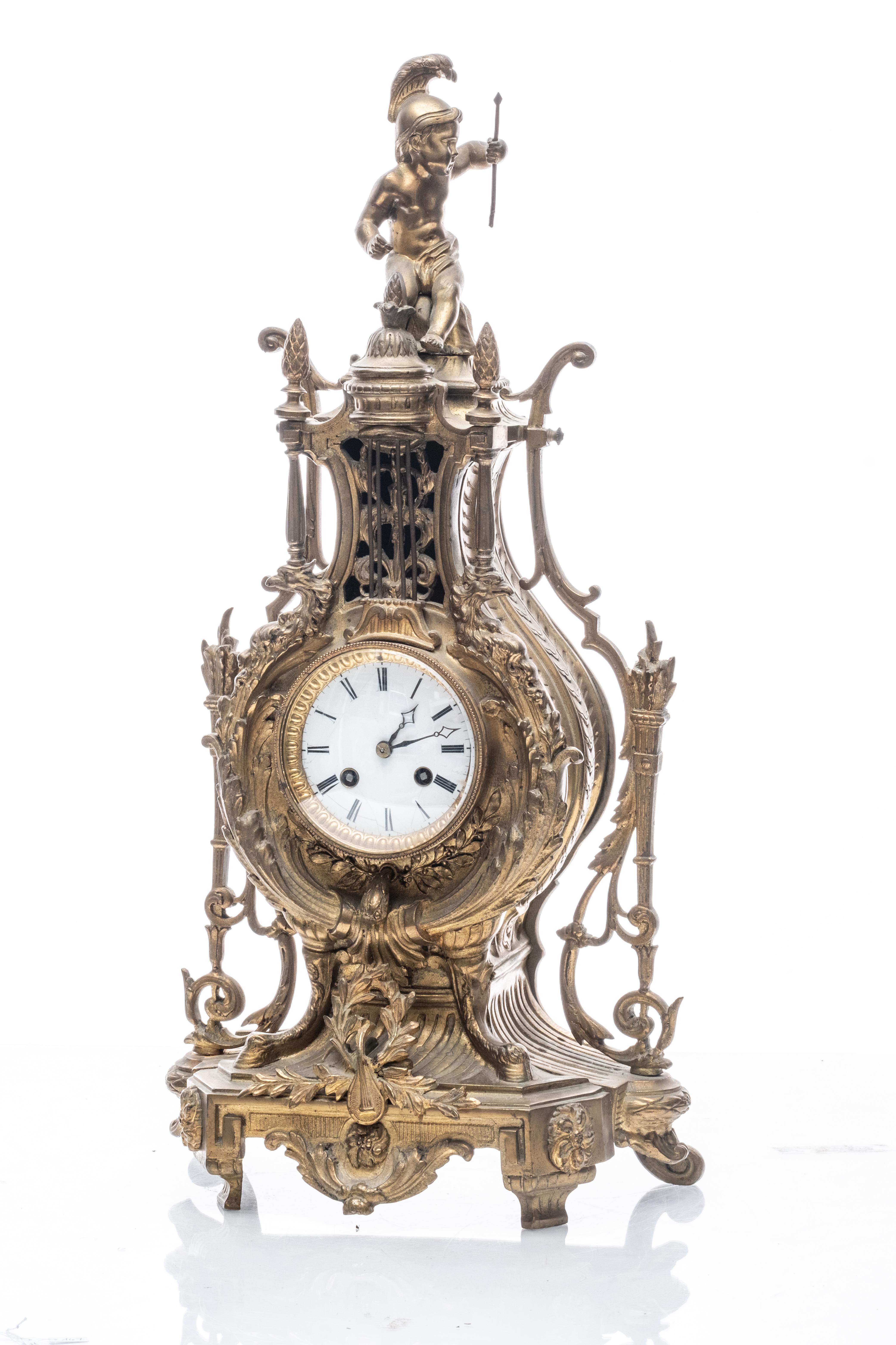 The bronze mantel clock is designed in a lyre form, featuring intricate decorative elements typical of 19th-century craftsmanship. It is surmounted by a cupid figure. The clock has a porcelain face with Roman numerals and ornate hands and comes with