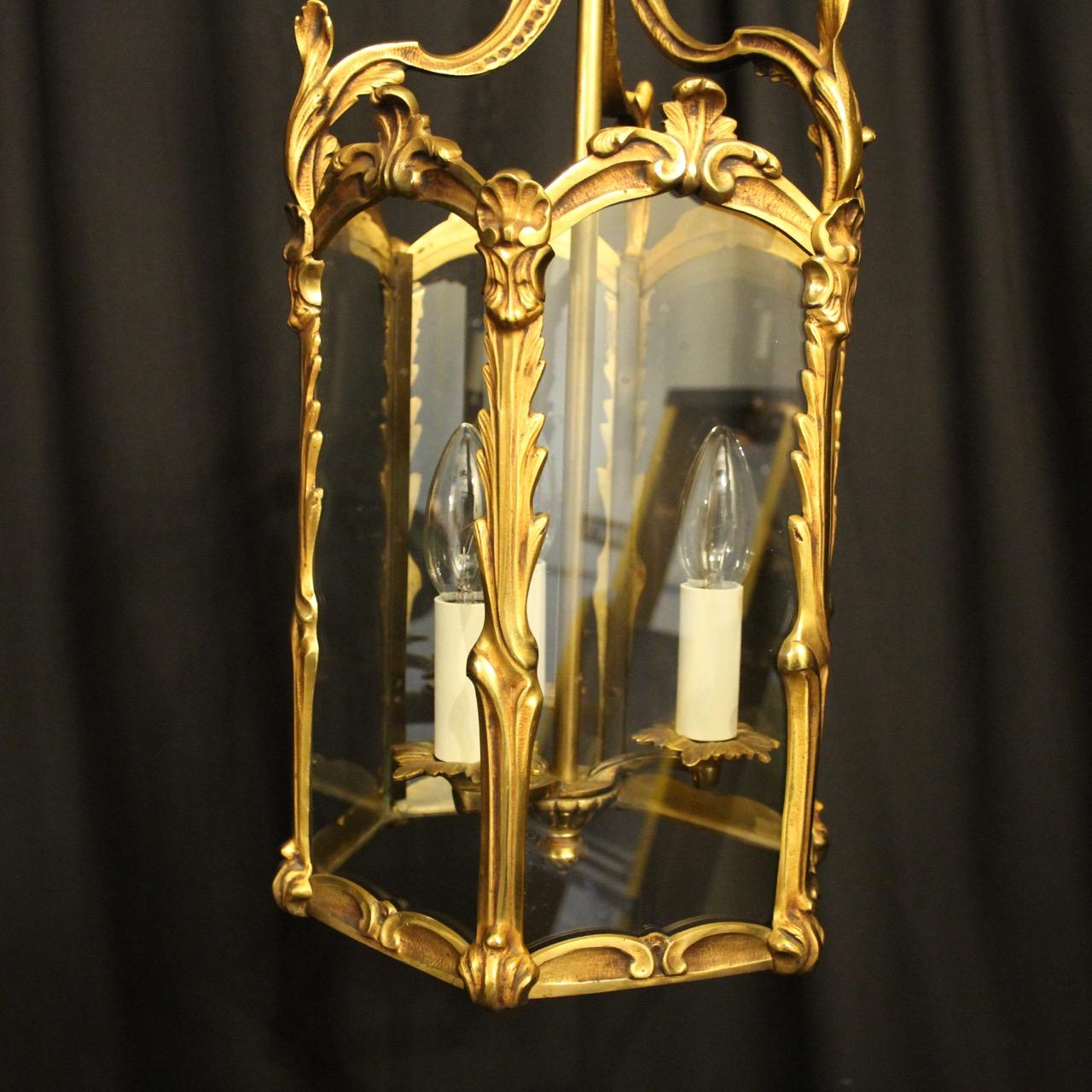 A French gilded bronze triple light hexagonal antique hall lantern, the six glass panels held within an ornate scrolling framework with three light fittings and having decorative scrolled central cartouche leaf embellishments, with leaf side panels