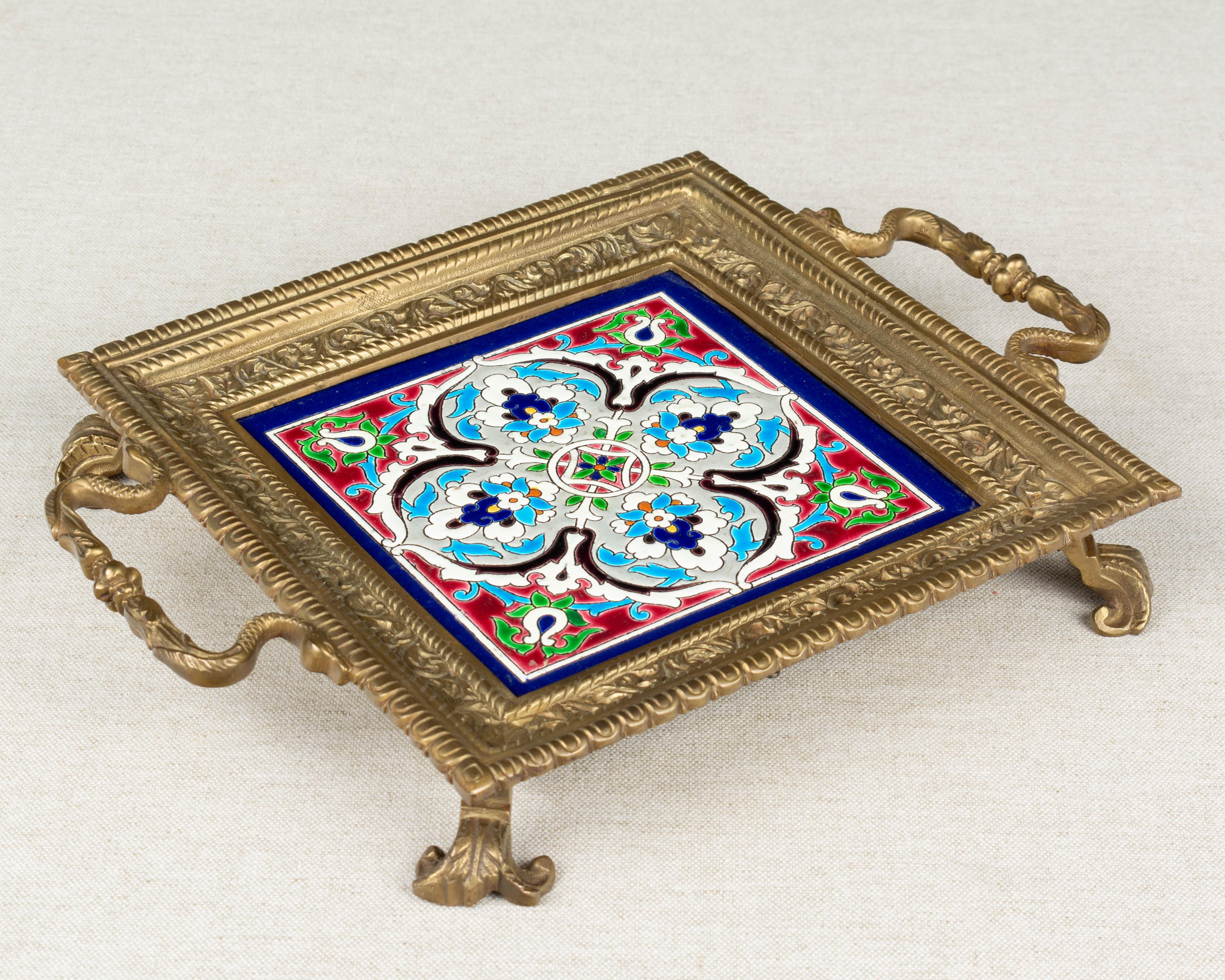 A French bronze trivet inset with a Longwy ceramic Persian style tile at the center. Vibrant hand painted cloisonné enamel in blue, red, green, yellow, orange and black on white and pale gray ground. Nicely detailed casting with handles at either
