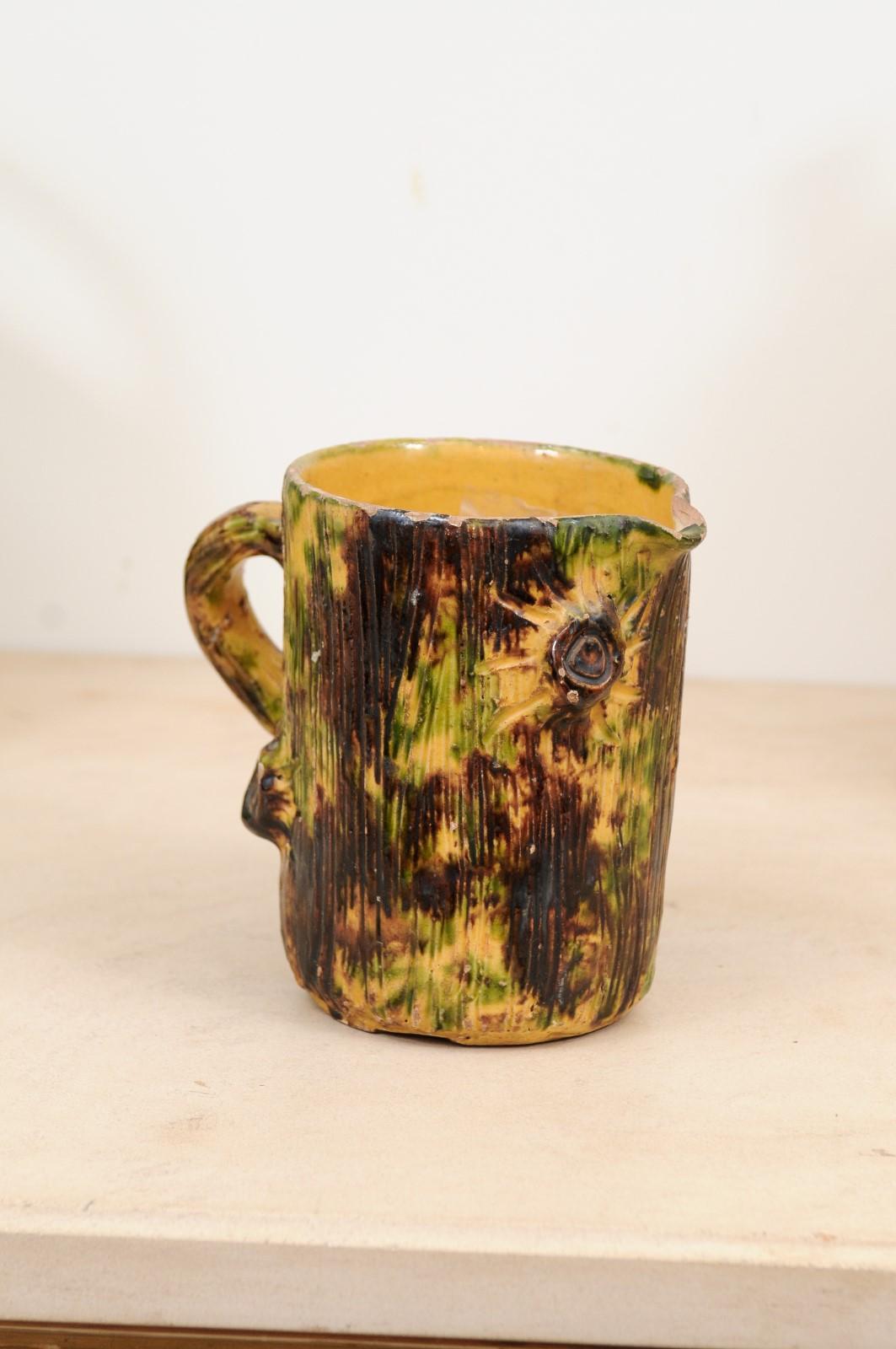 A French glazed pottery pitcher from the 19th century, with textured finish and floral motifs. Created in France during the 19th century, this rustic pitcher features a brown textured glaze adorned with green and yellow accents and a floral motif.