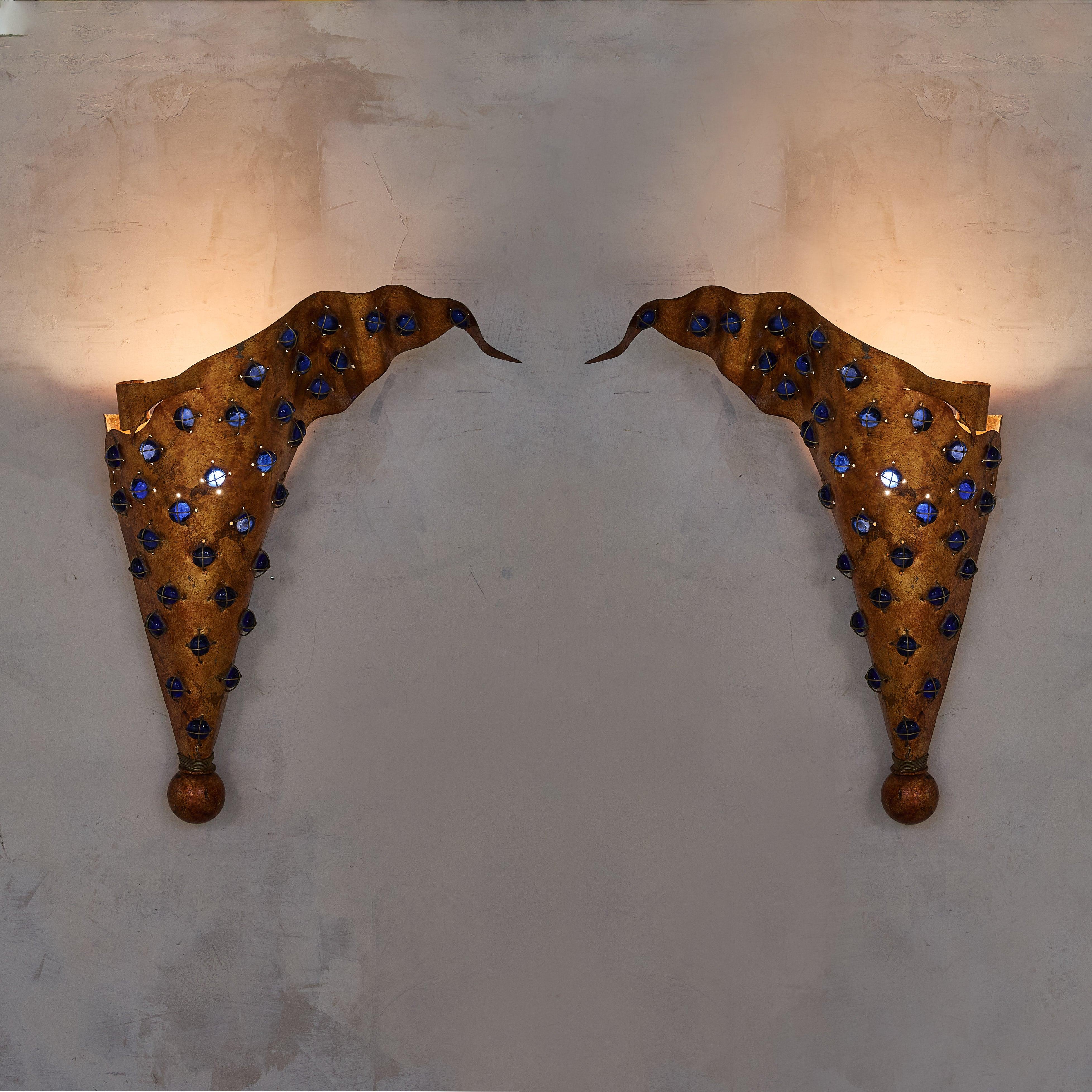 A pair of 1970s French wall sconces, crafted in a conical shape with fascinating, free-flowing edges and an opulent brushed gold finish. Accented with vivid blue glass cabochons and spherical bases, their sculptural forms emit a warm, atmospheric