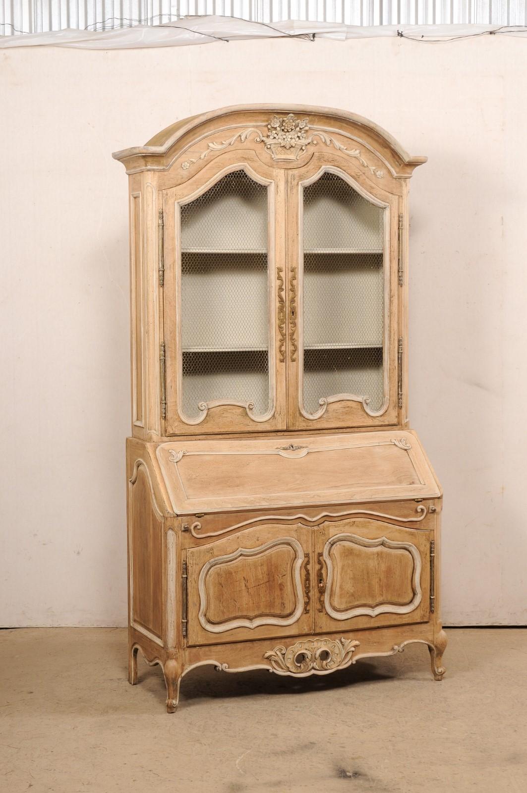 A tall French Louis XV style carved and bleached wood display and storage cabinet with secretary. This vintage cabinet from France is topped with a thickly carved arched bonnet pediment, which perfectly frames the corbeilles de fleurs (basket with