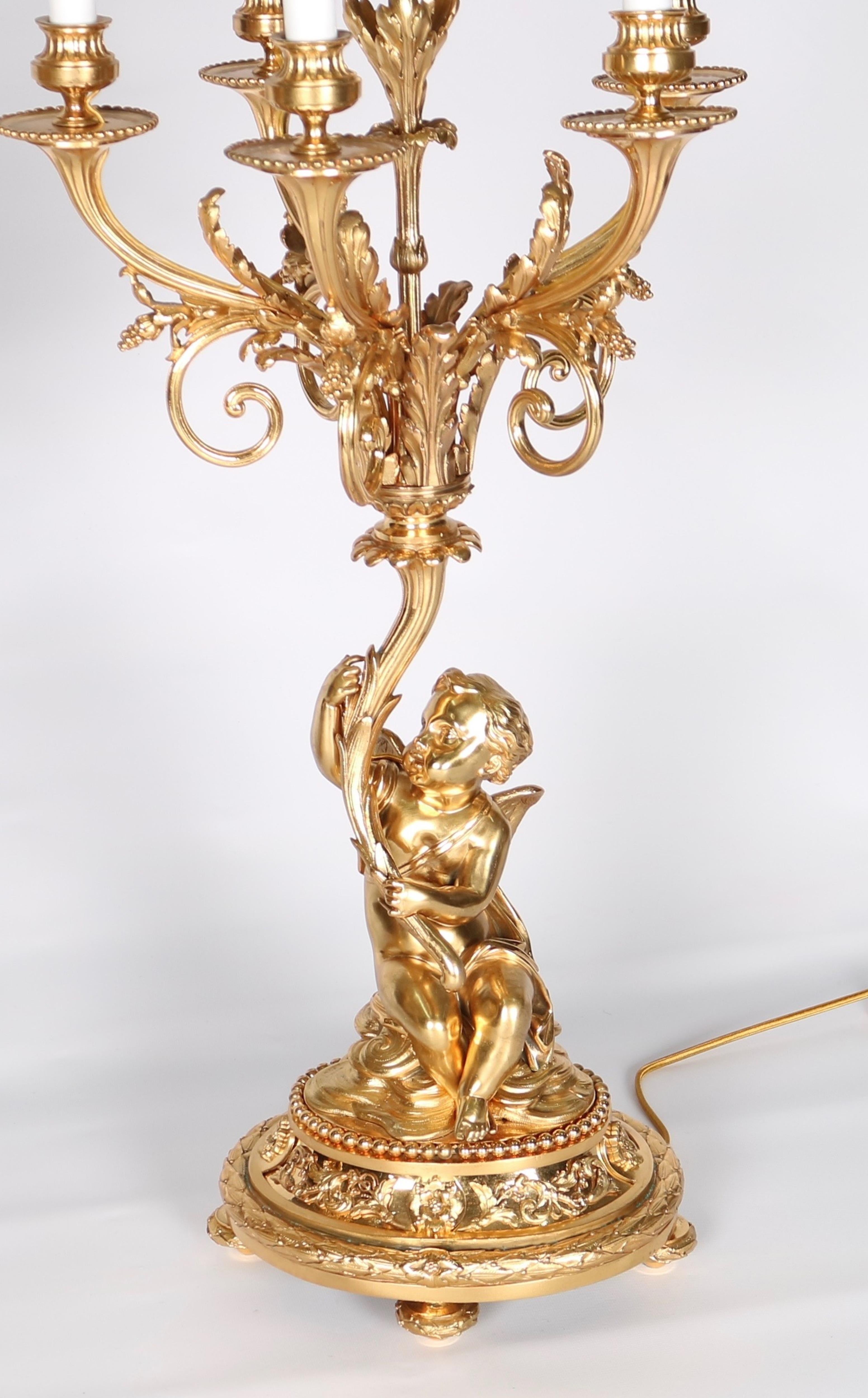 French candelabra lamps in gilt bronze with putti. Each lamp stands on an oval base with a seated putti holding an arrangement of curved acanthus leaves ending in six arms. Crafted in the 18th century, the pair have been newly rewired. Despite wear