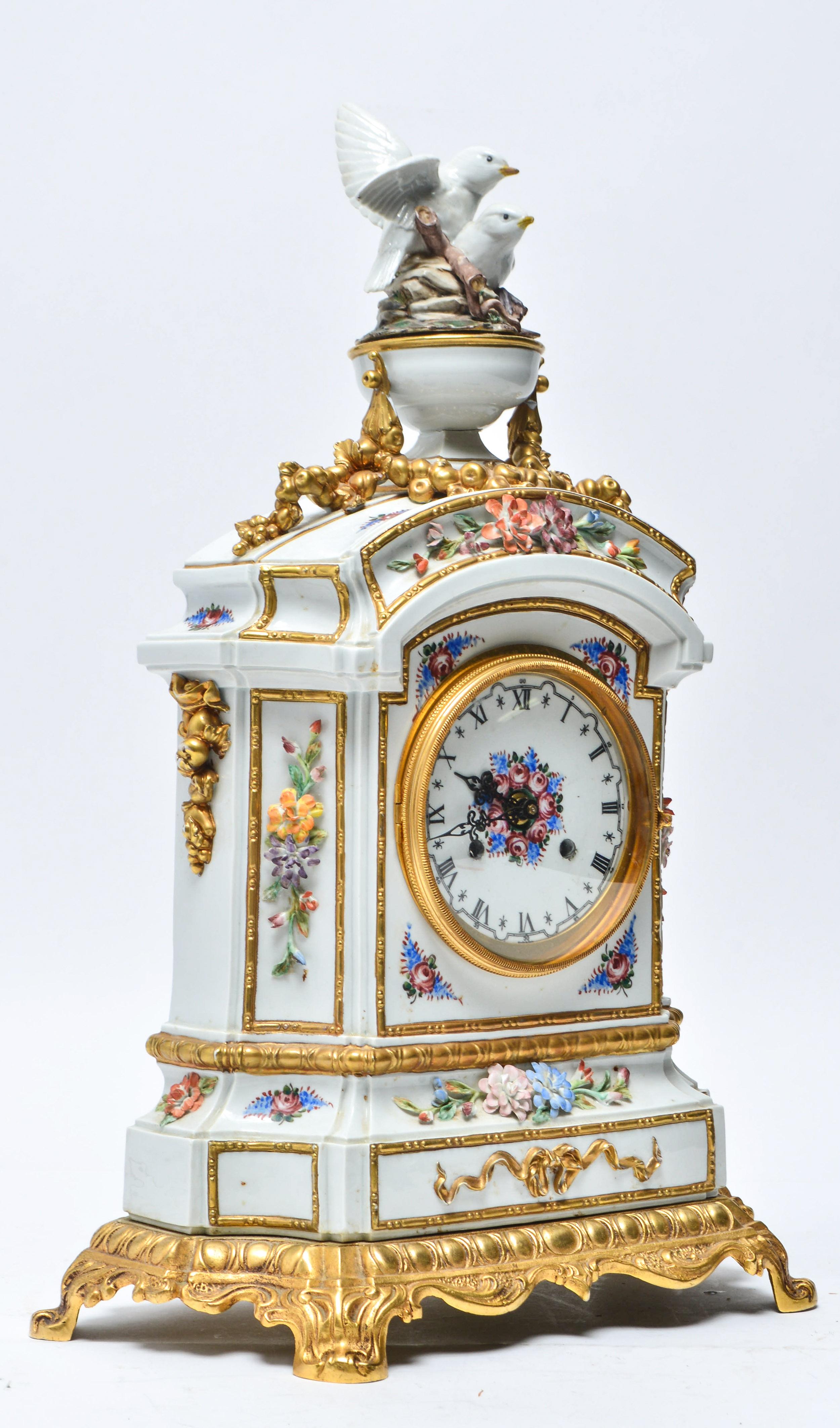French Carpie hand painted mantel clock with gilt-trim porcelain and mounted on a fitted shaped brass base. The piece has a finial of decorative doves and applied flowers. The dial has black Roman numerals. Marked: 