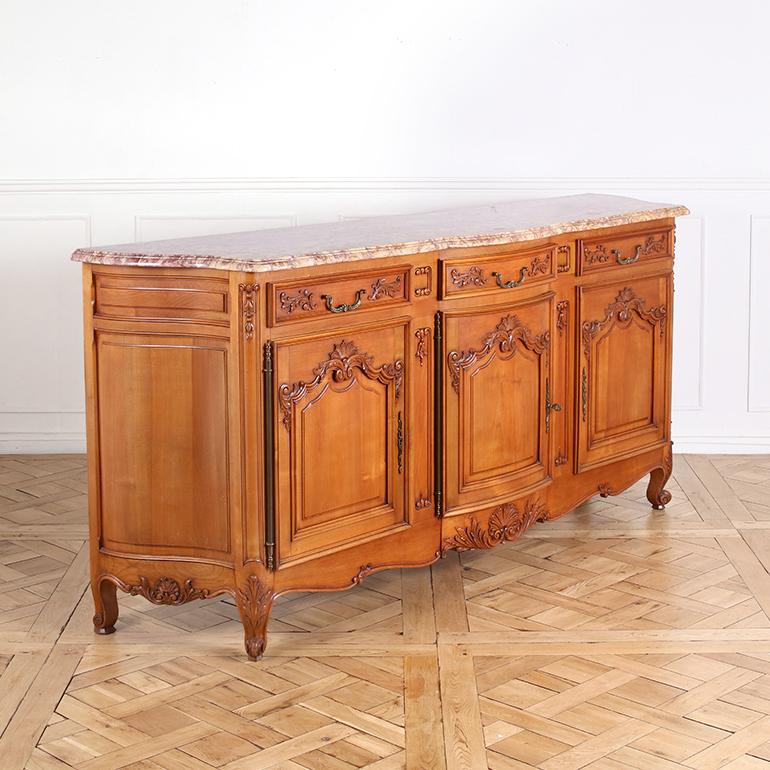 Questo elegante buffet francese in stile Luigi XV è realizzato in legno di ciliegio riccamente venato e sormontato da un marmo sagomato e smussato. La sua silhouette dolcemente arcuata e i dettagli finemente intagliati riflettono gli elementi di