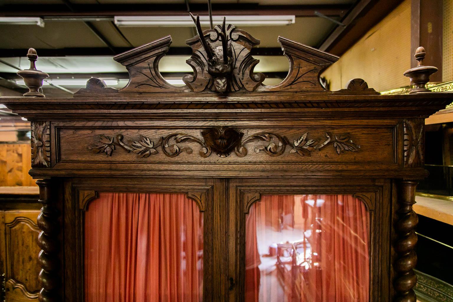 The cornice of this oak cupboard has a carved stag head with leaf carvings in a central cartouche in the frieze. The side stiles in the upper and lower halves are barley twist. The upper case is supported by stylized winged lions. The drawer handles
