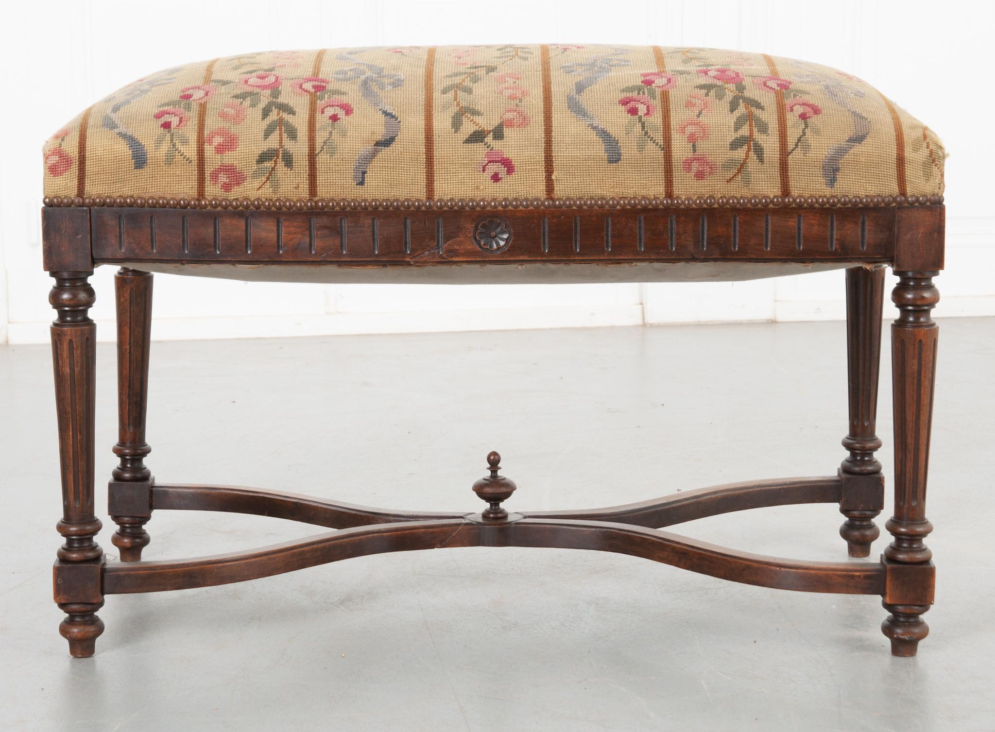A wonderful walnut bench from 19th century France! The needlepoint upholstery is slightly worn but retains its colorful hand-crafted details; pink roses on the vine and blue ribbon tied in bows. The fabric is secured by a perimeter of decorative