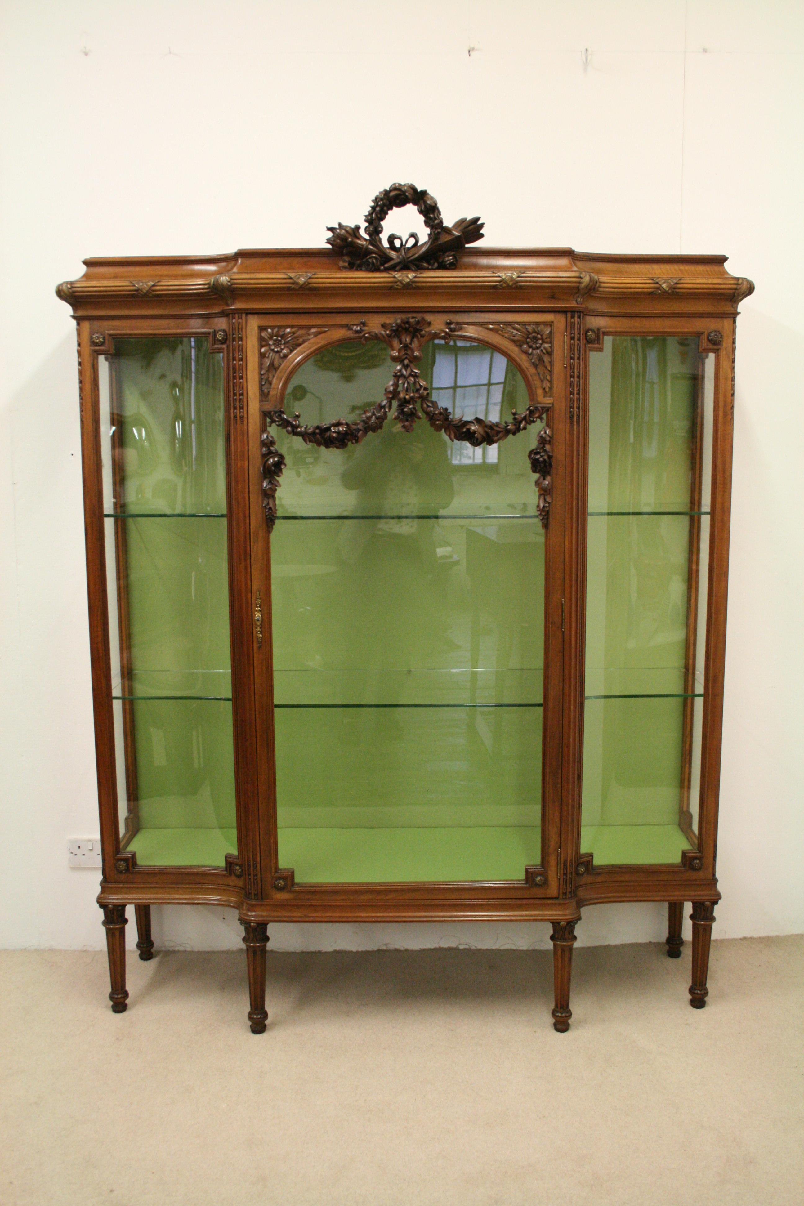 French carved walnut display cabinet. Mounted with a carved wreath of flowers and carved burning torches, swags and tails, and ribbon. The mounted cornice has a ribbed and cross over ribbon and gilded carvings in the corners, and beneath is a full