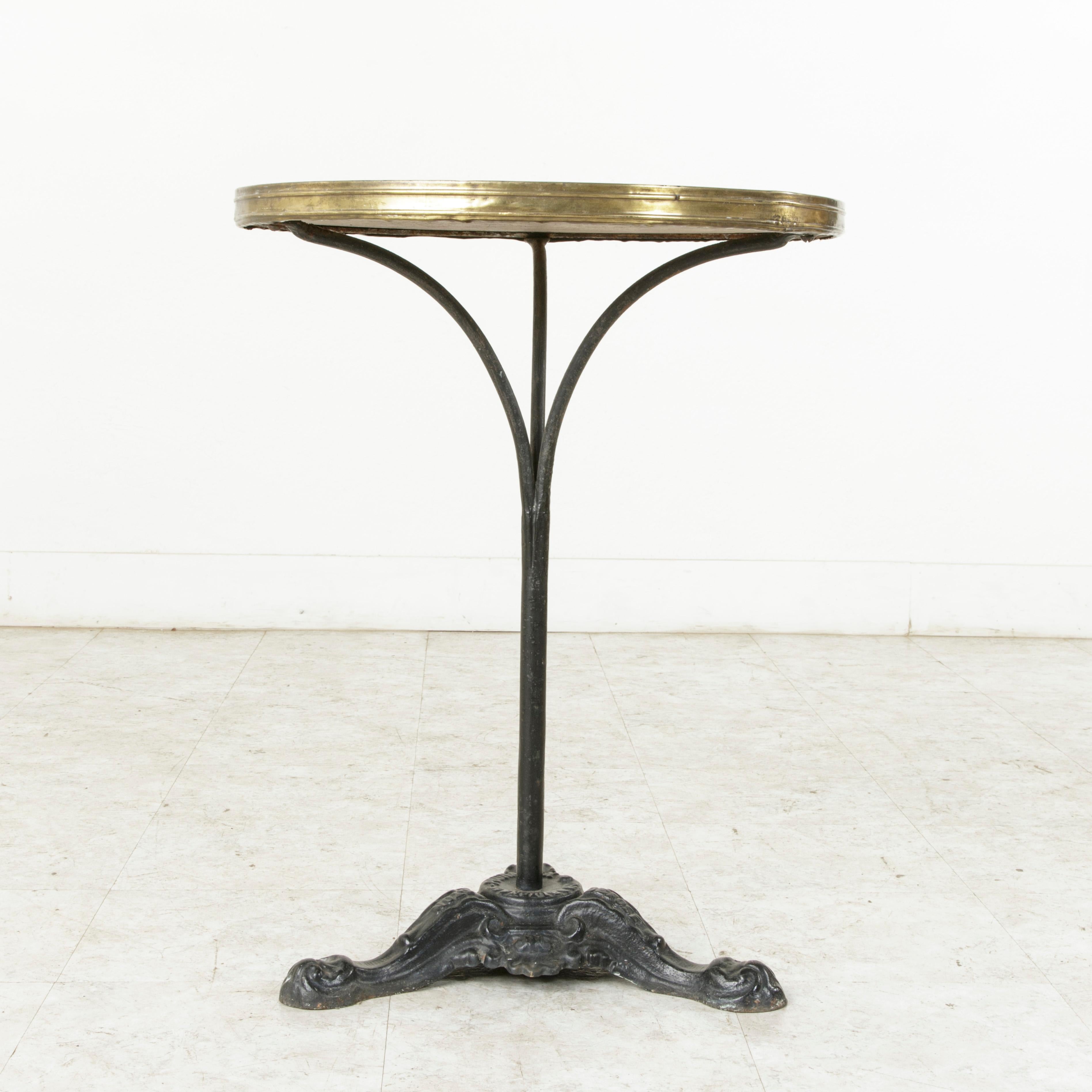 This French iron bistro table from the turn of the 20th century features its original white marble top surrounded with brass banding. Three arms support the top and extend down to a central pillar. Its enameled iron base is decorated with a shell