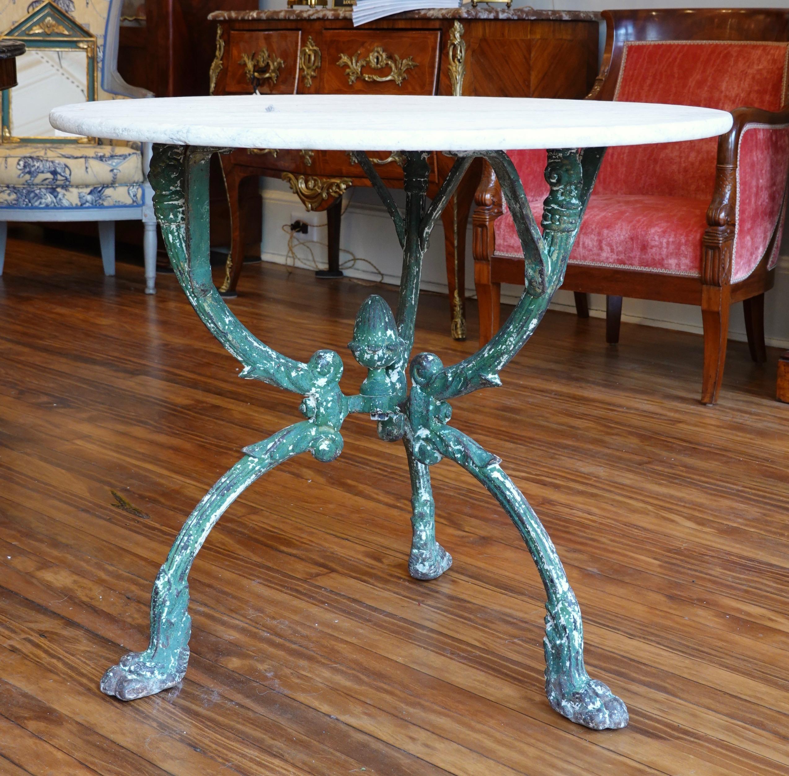 This French cast iron garden table has an old green painted finish and features a highly decorate tripod base with paw feet, an acorn finial, and the top of each leg terminating with a caryatid's head. The white Carrera marble top with grey veining