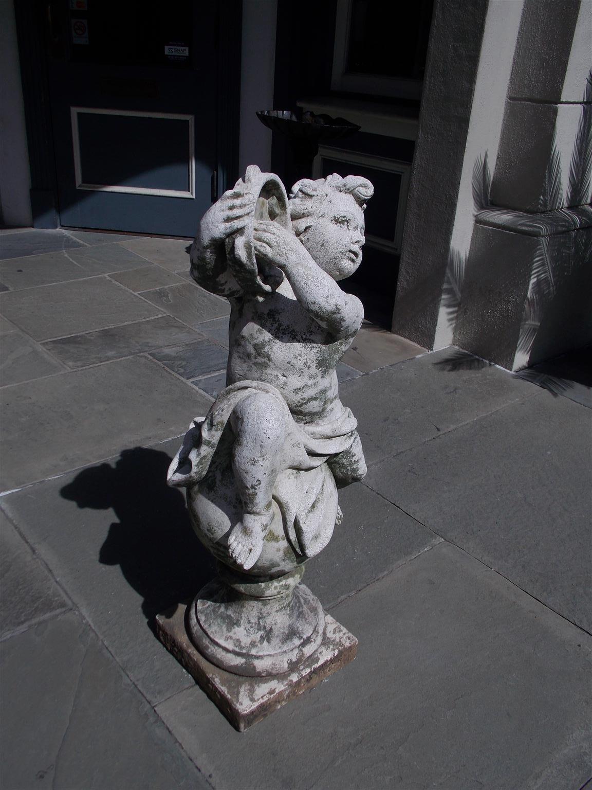 French cast stone musical tambourine putti garden ornament seated on a sphere form with a squared plinth, and decorative sash. Mid-19th century.