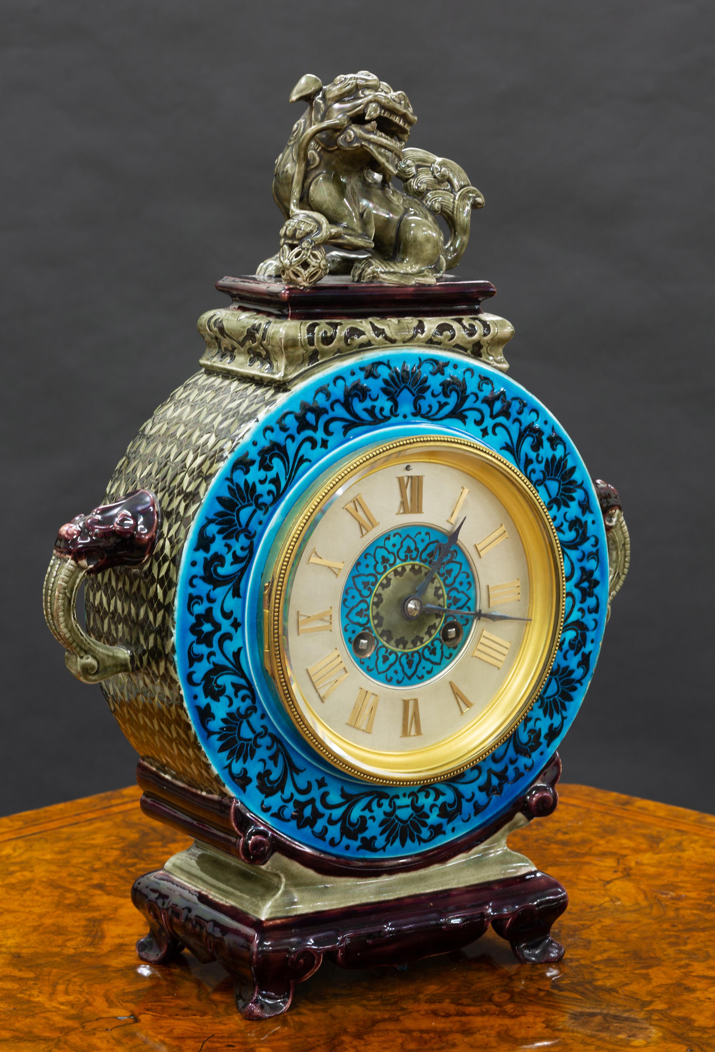 French ceramic clock with Chinese decoration standing on outswept feet with purple and turquoise glazed decoration and lions head ‘carrying’ handles surmounted by a Foo dog resting his foot on a lattice work ball.

Raised beaded decorated gilded