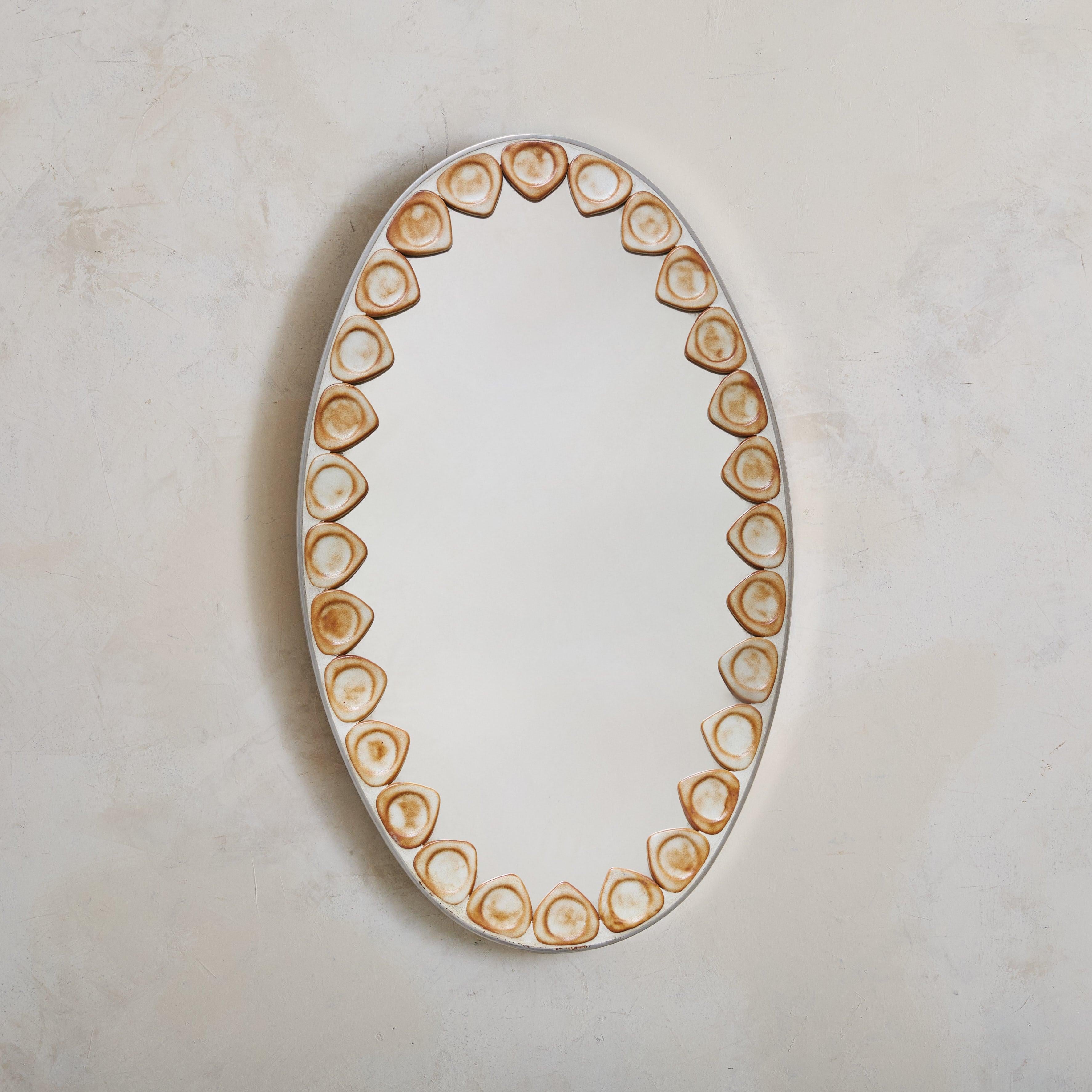 A French artisan-crafted ceramic wall mirror from the 1960s. The oval frame, made of silver metal, is accented with organic ceramic embossments, each featuring matte ivory and burnt sienna glazes. This intricate detailing creates a captivating play