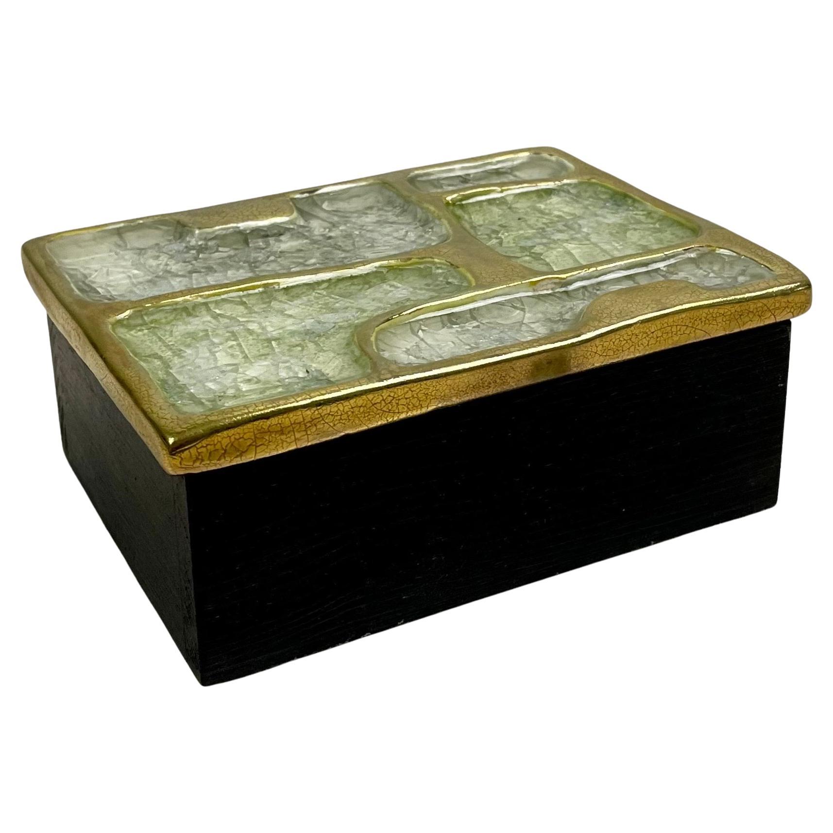 Boîte à secrets, boîte à bijoux, boîte de rangement, rangement rectangulaire avec couvercle en céramique émaillée dans les tons or, blanc et pellucide et base en bois, par Mithé Espelt.
La boîte est décorée de formes géométriques. Cette boîte donne