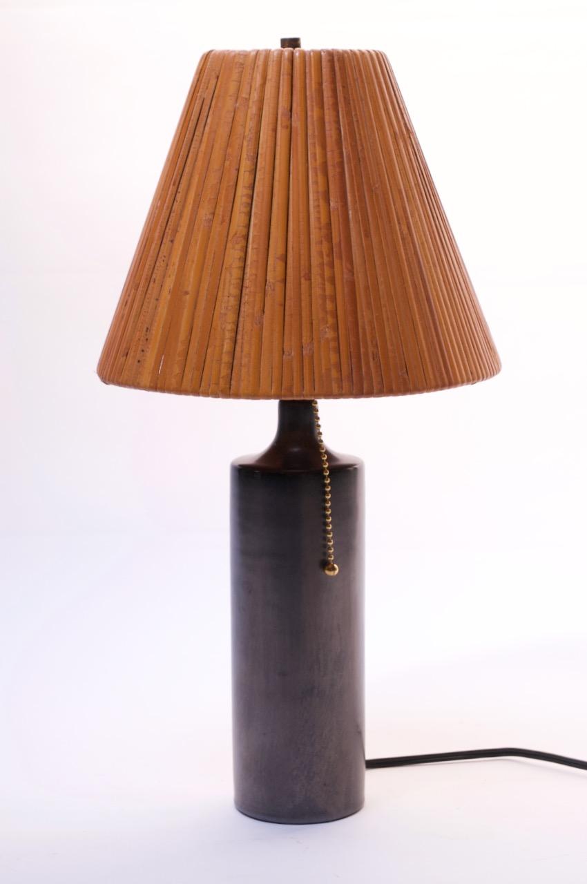 Petite ceramic table lamp by Georges Jouve in a metallic graphite-bronze matte finish, circa 1960. Socket and pull string have been added in the rewiring process. Additionally, the vintage rattan shade has been paired with the piece (it is included,