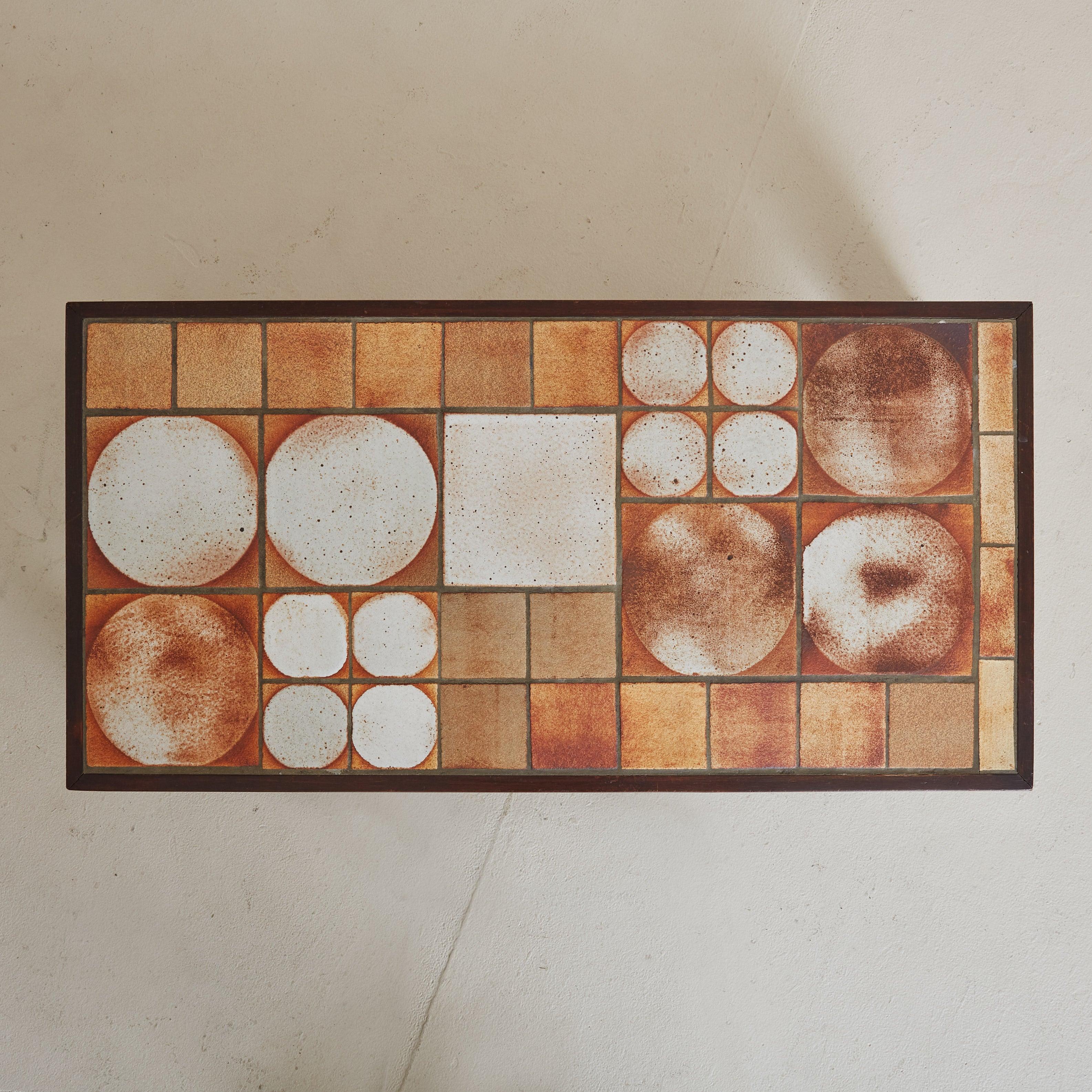 A 1950s low coffee table, shaped by the skilled hands of an artisan from La Borne, France. The rich wooden frame, grounded by four block legs, supports a mosaic of ceramic tiles. In warm, earthy tones of sand and terracotta, the tiles create a
