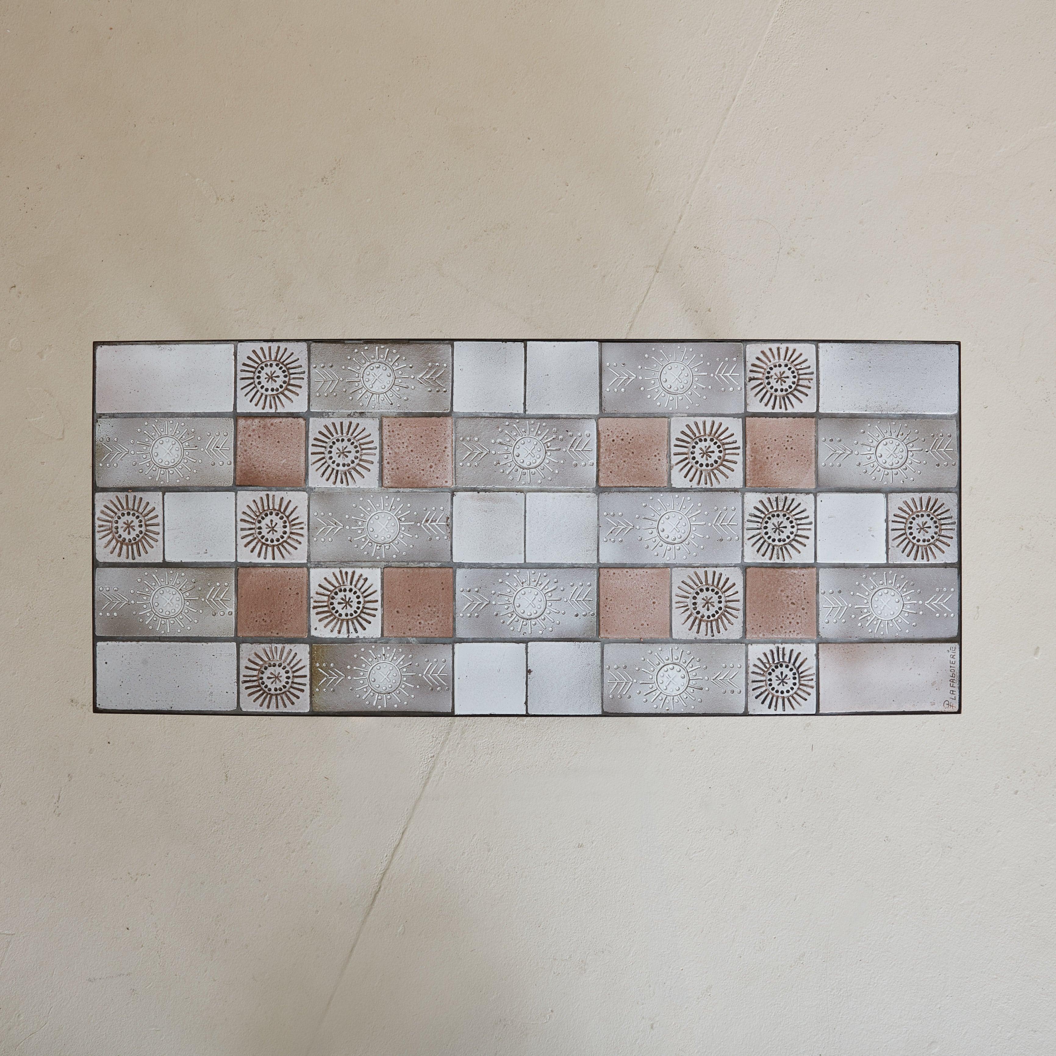 A 1960s coffee table, handcrafted by a French artisan and signed ‘La Faboterie’. The rectangular top is a mosaic of hand-painted ceramic tiles from Vallauris, France, displaying a muted palette of soft gray, white, and faded red, with a few