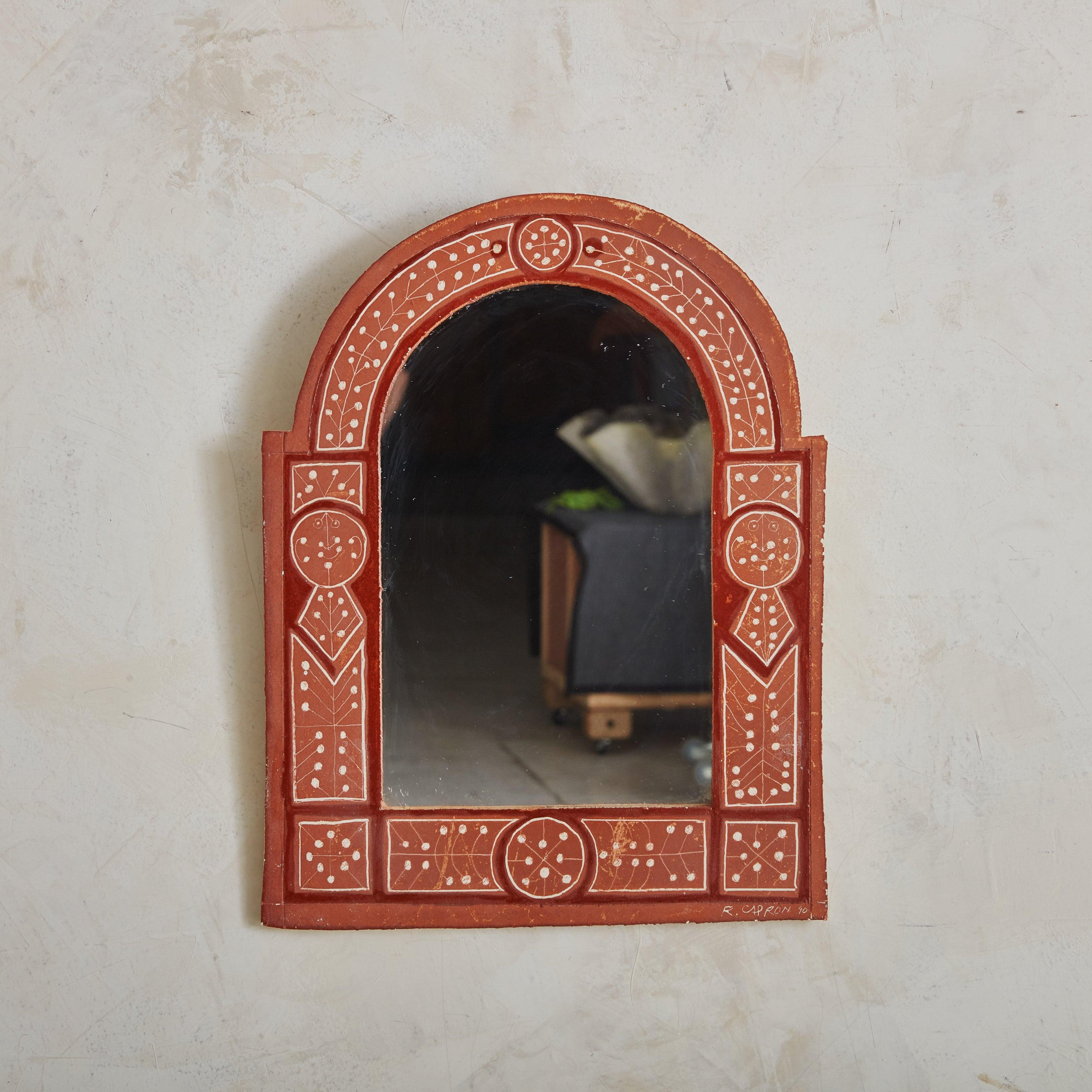 Petit miroir mural des années 1950 du céramiste français Roger Capron. Le cadre en céramique de couleur terre cuite, peint à la main avec des motifs géométriques et floraux, capture l'essence de l'art populaire méditerranéen. Sa silhouette arquée,