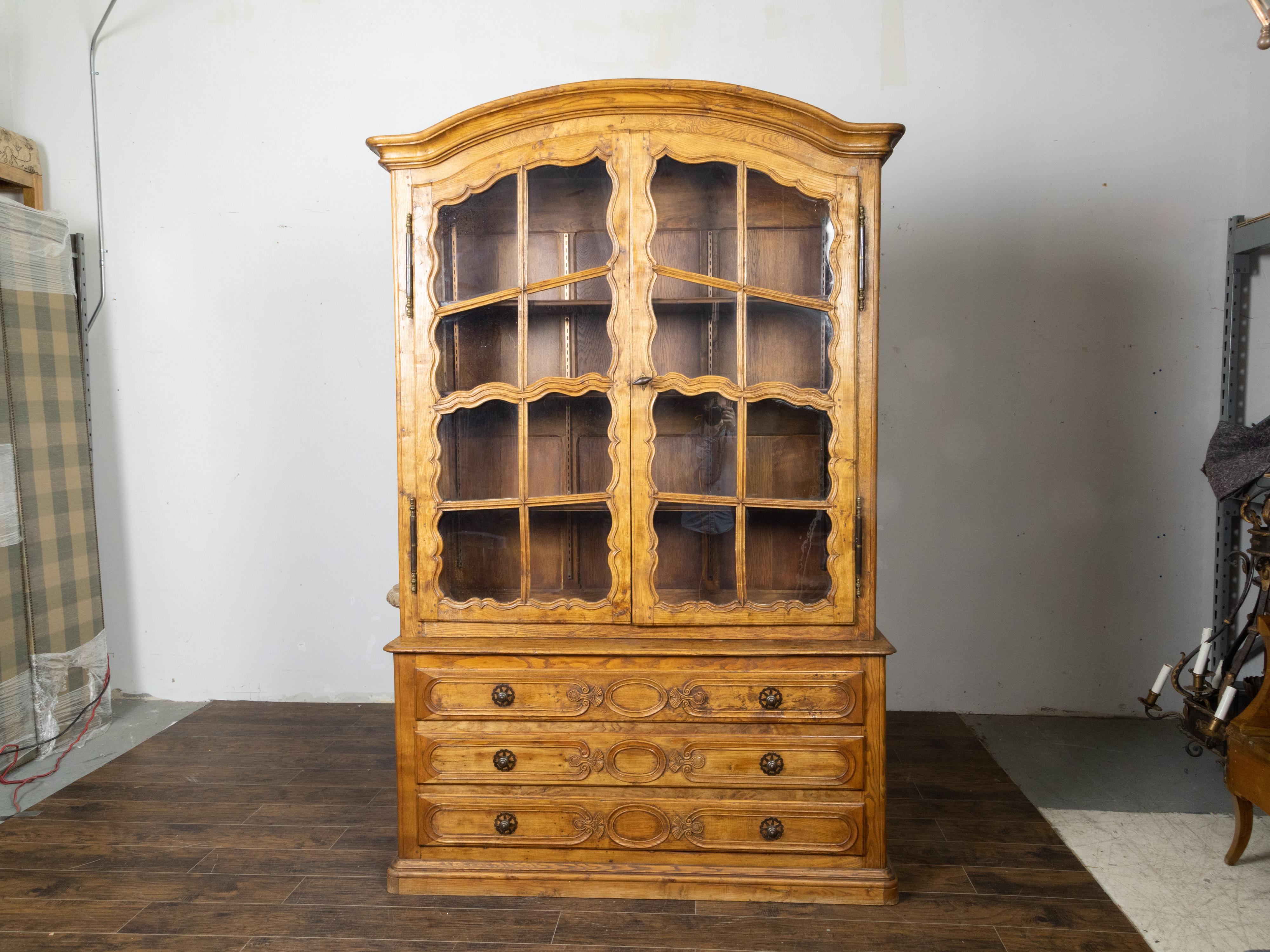 A French elm vitrine cabinet from the 19th century, with chapeau de gendarme top, glass doors, carved décor and three drawers. Created in France during the 19th century, this elm vitrine cabinet features a narrow upper section topped with a chapeau