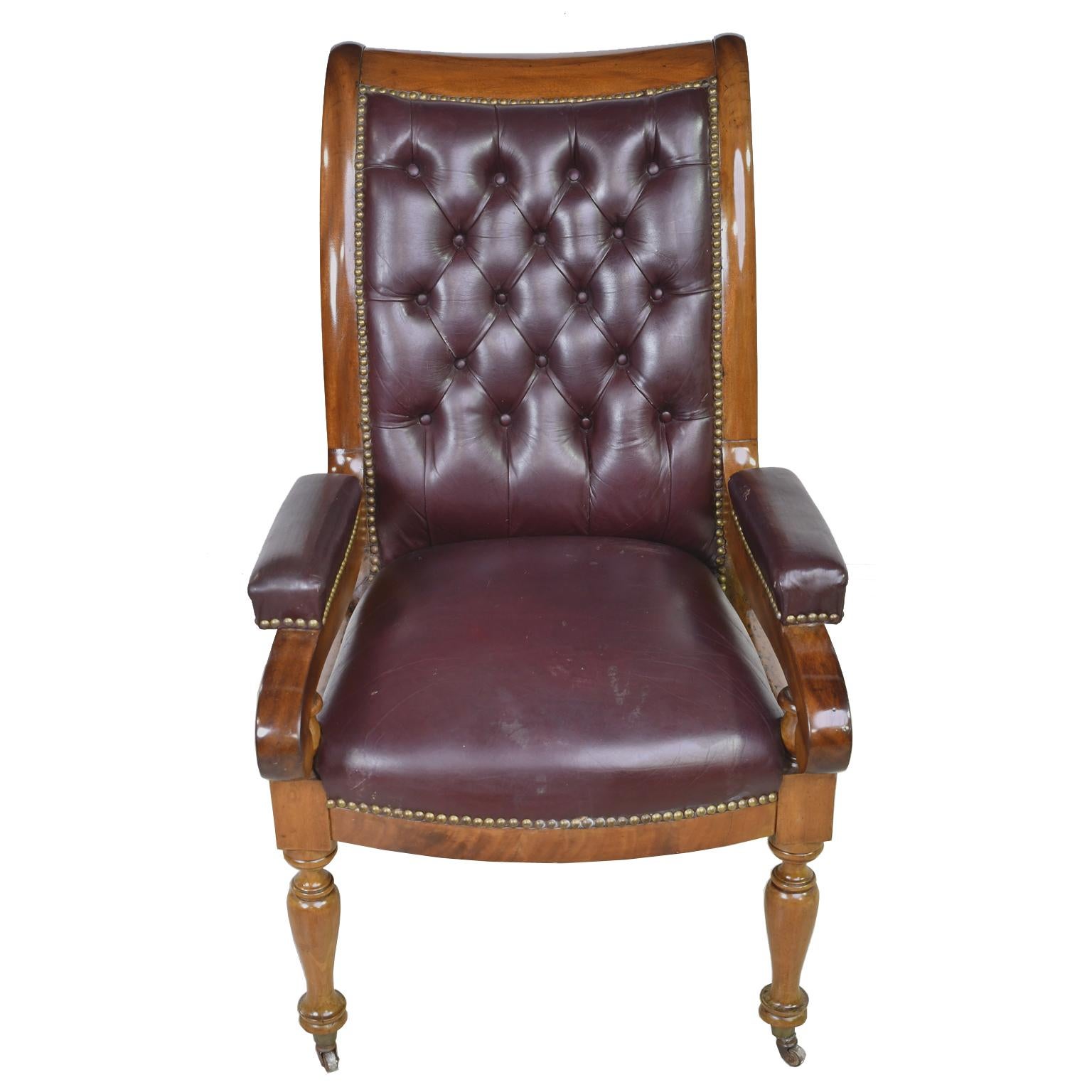 A fine Charles X desk chair in mahogany with scrolled arms, curved/ bowed seat, turned front legs on porcelain casters, and square saber, rear legs. France, circa 1820. The frame has a burgundy/brown- colored leather upholstery with decorative brass