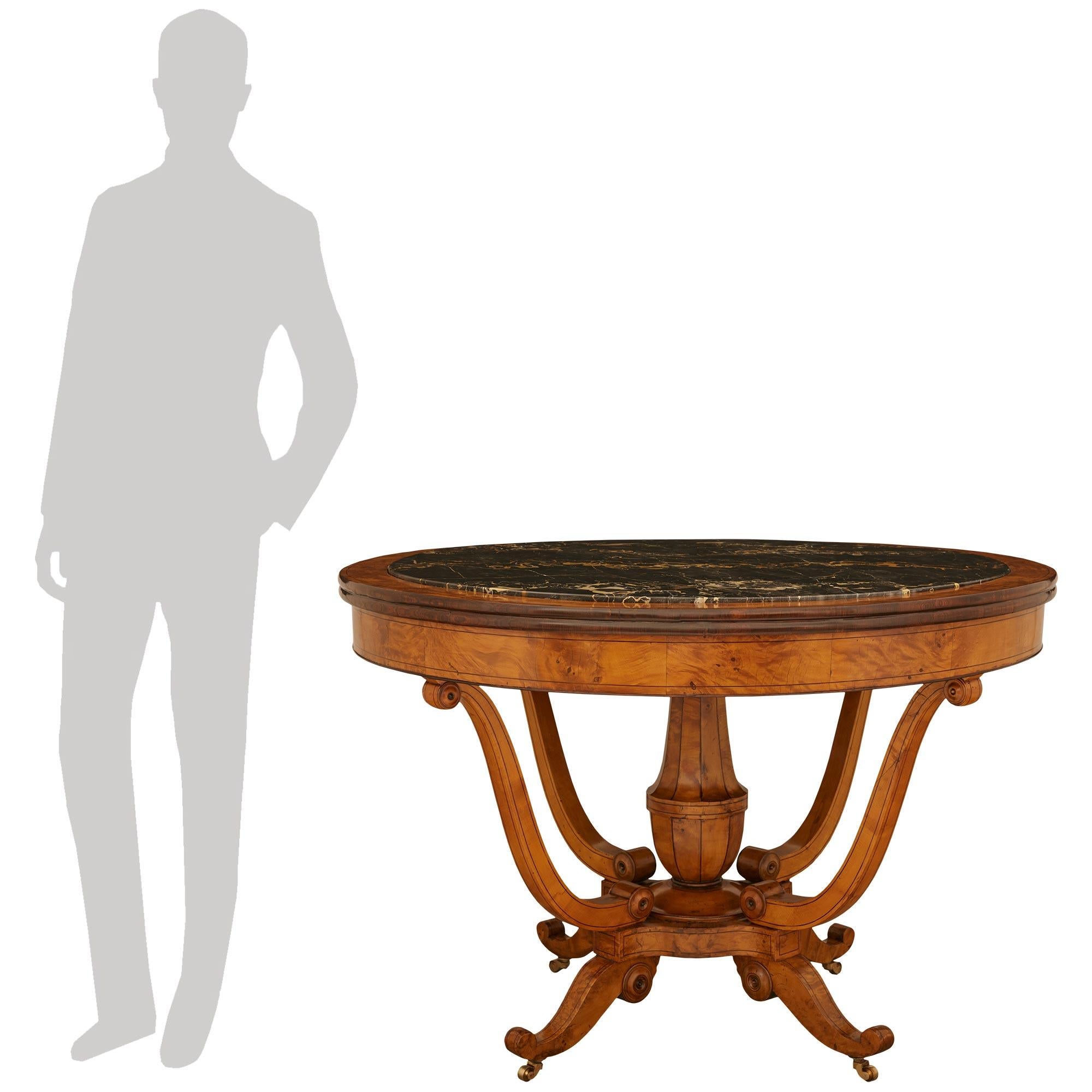Atractiva mesa de centro de madera de arce, abedul y mármol del periodo Charles X francés del siglo XIX. Esta hermosa mesa se eleva sobre cuatro ruedas de bronce que sostienen patas cortas de madera de arce con volutas que terminan en grandes