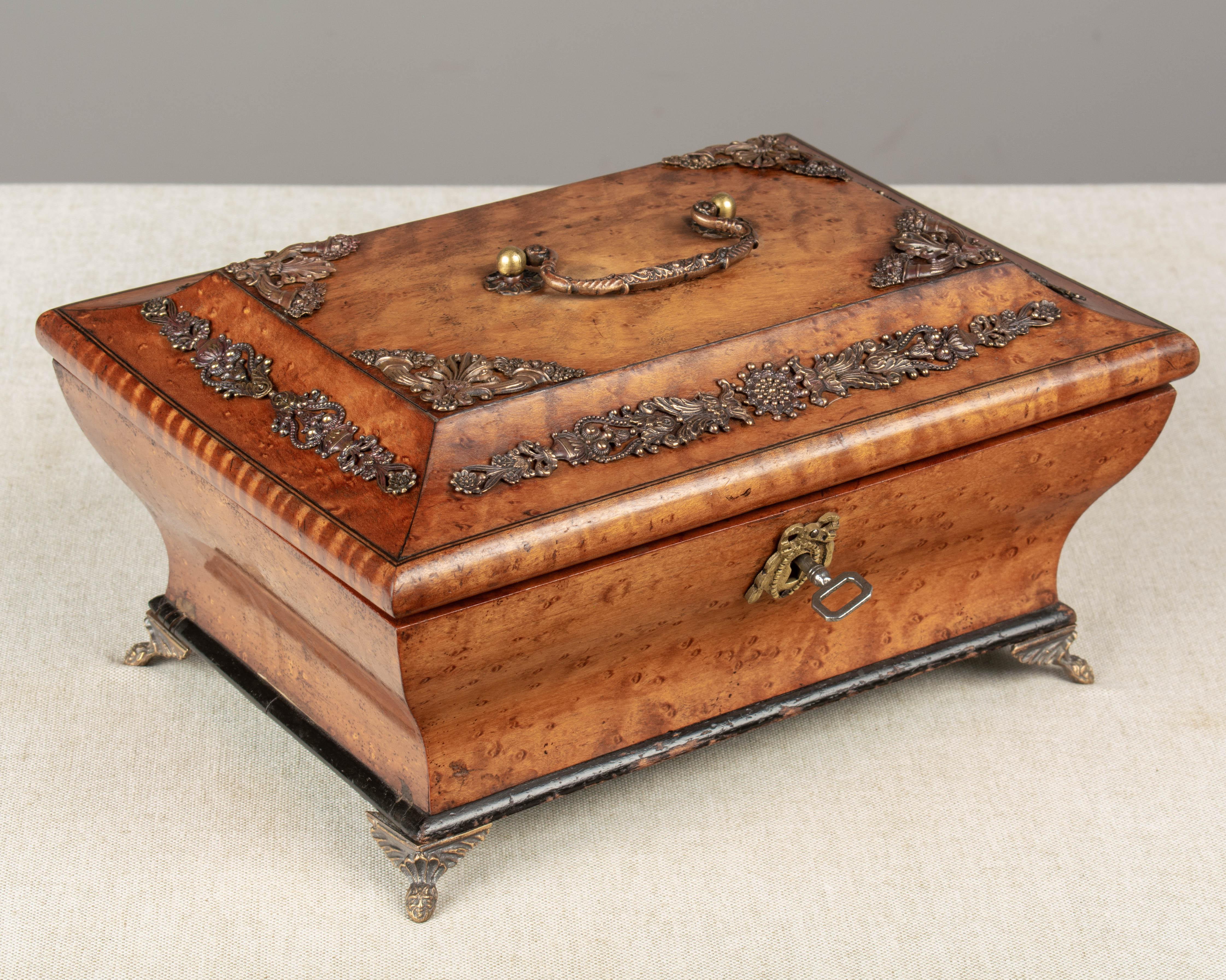 An early 20th century French Charles X style jewelry or sewing box made of amboyna wood veneer with cast brass decoration and handle. Interior is lined in blue moiré fabric with mirror inside the lid. Raised on small brass feet. Black painted trim