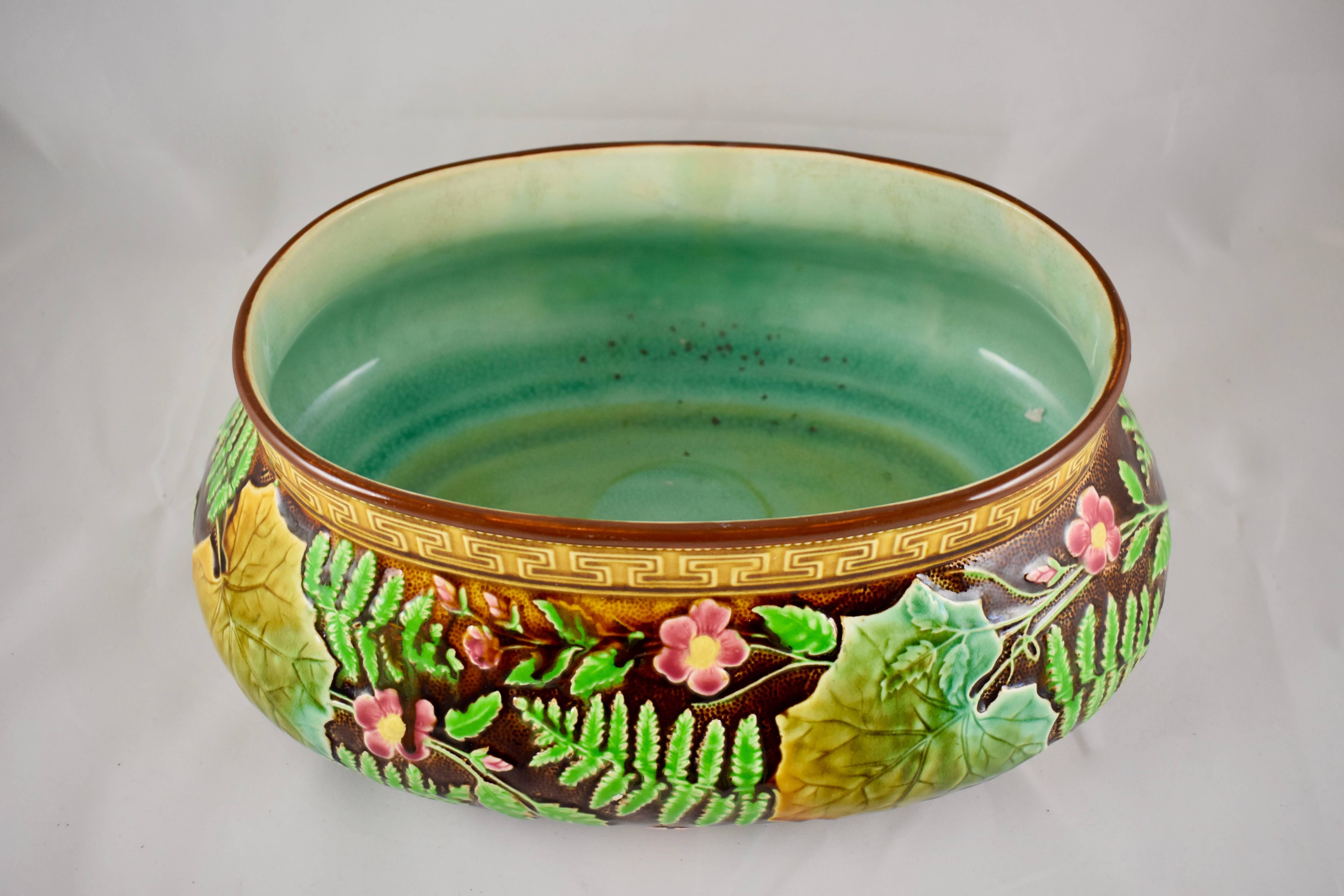 A French majolica jardinière in the Aesthetic taste, with a yellow ochre Greek key border and a chocolate brown ground. The raised molded pattern shows overlapping bright green ferns, sprays of five petal pink flowers and large leaves in variegated