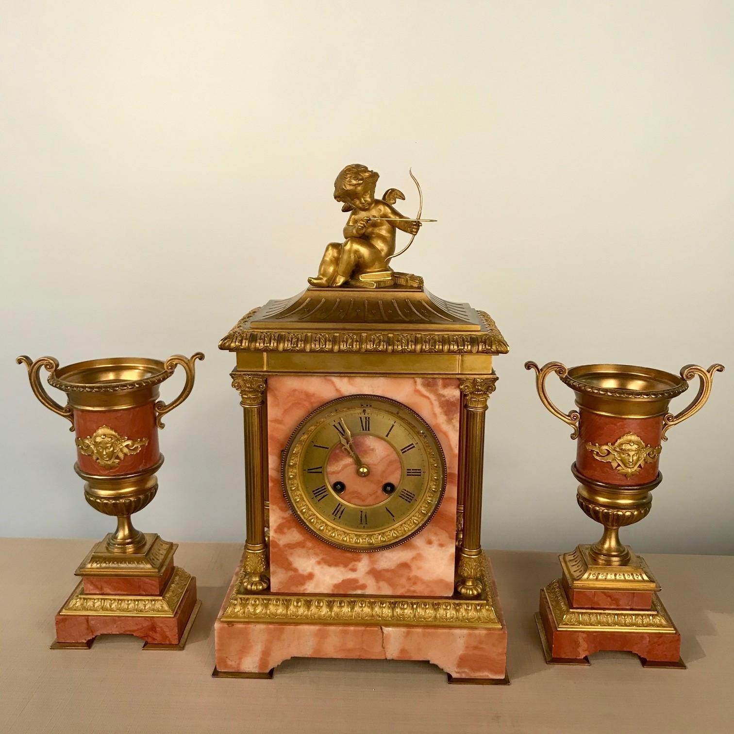 This set is a particularly fine example of this sort mantle ornament; The clock, surmounted by a Cupid with bow and arrow, is flanked by a pair of Campana shaped urns. The marble is an exquisite salmon colour, (most unusual) the half fluted urns are