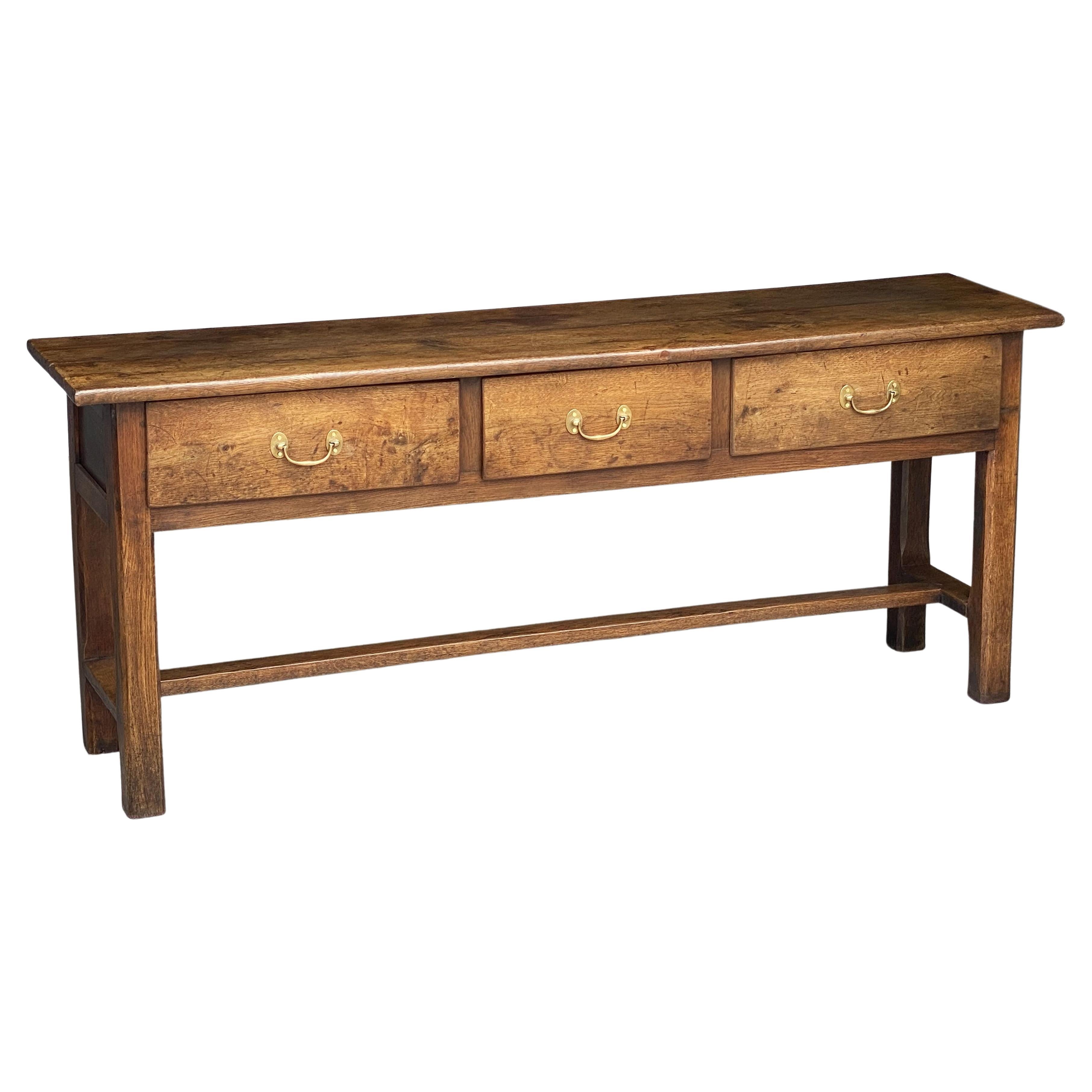 A handsome French console sideboard or serving table of chestnut wood featuring a plank top over a paneled frieze with three drawers, each with brass hardware pulls, and resting on a chamfered leg stretcher base.