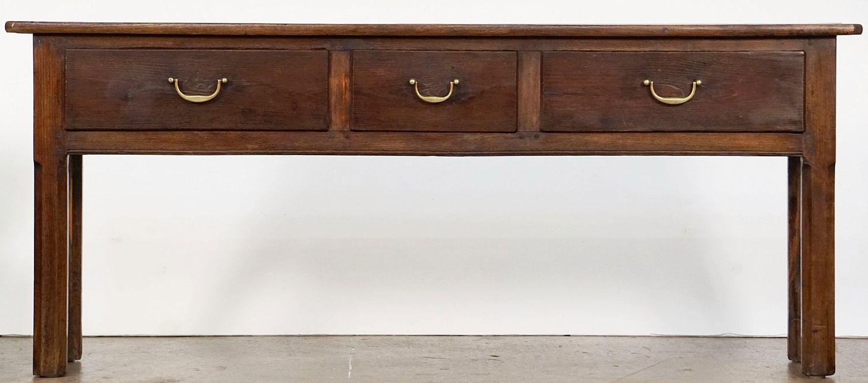 A handsome French console sideboard or serving table of patinated chestnut wood, featuring a plank top over a paneled frieze with three drawers, each with brass hardware pulls, and resting on four chamfered legs.